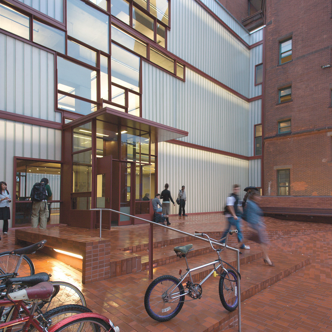 Image of a modern academic building entrance with large glass and metal panels. The design features a combination of brick and metal accents, with steps leading to the main door. A bike is parked in the foreground, and students are seen walking in and out, adding a dynamic feel to the space. The architecture reflects a blend of contemporary and industrial styles, with a focus on clean lines and open spaces.