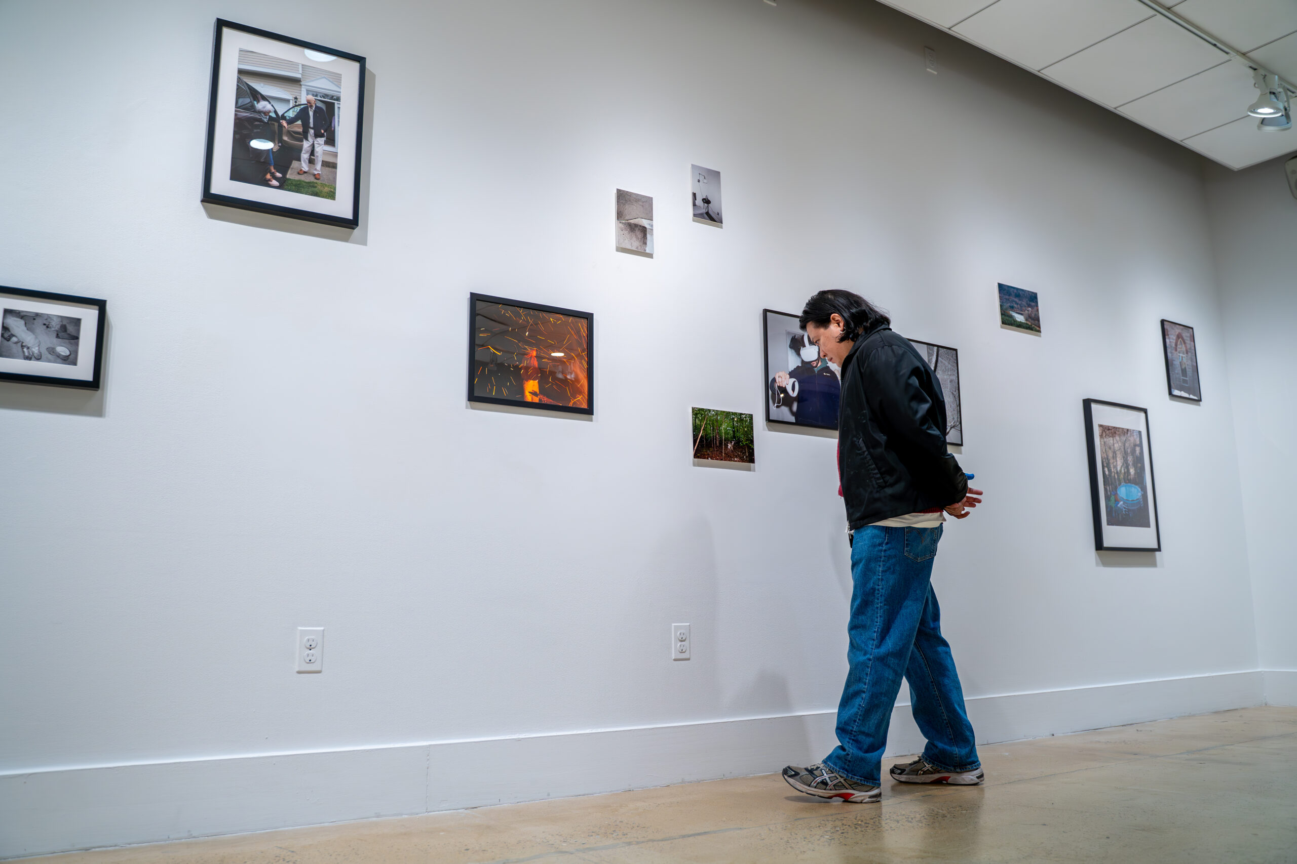 A group of people is scattered throughout an art gallery space, some standing near the artworks while others sit on a bench in the center. The white walls display framed photographs, and the space is brightly lit with overhead lights.