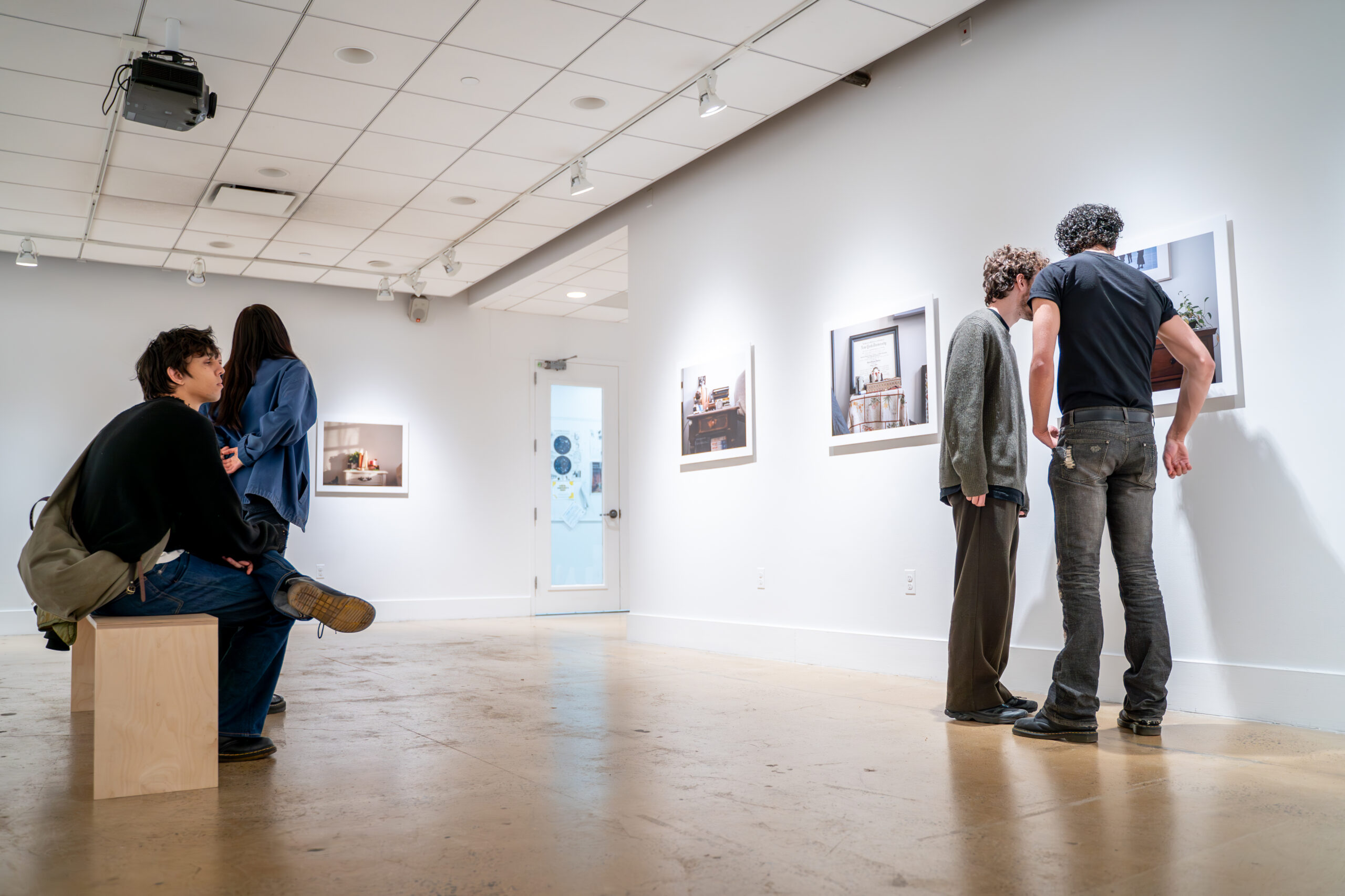 Interior of an art gallery with people observing framed photographic artworks on white walls. A man in black with an apron stands on the right, while others interact in the background. The space is brightly lit with overhead lights and has a minimalistic aesthetic.