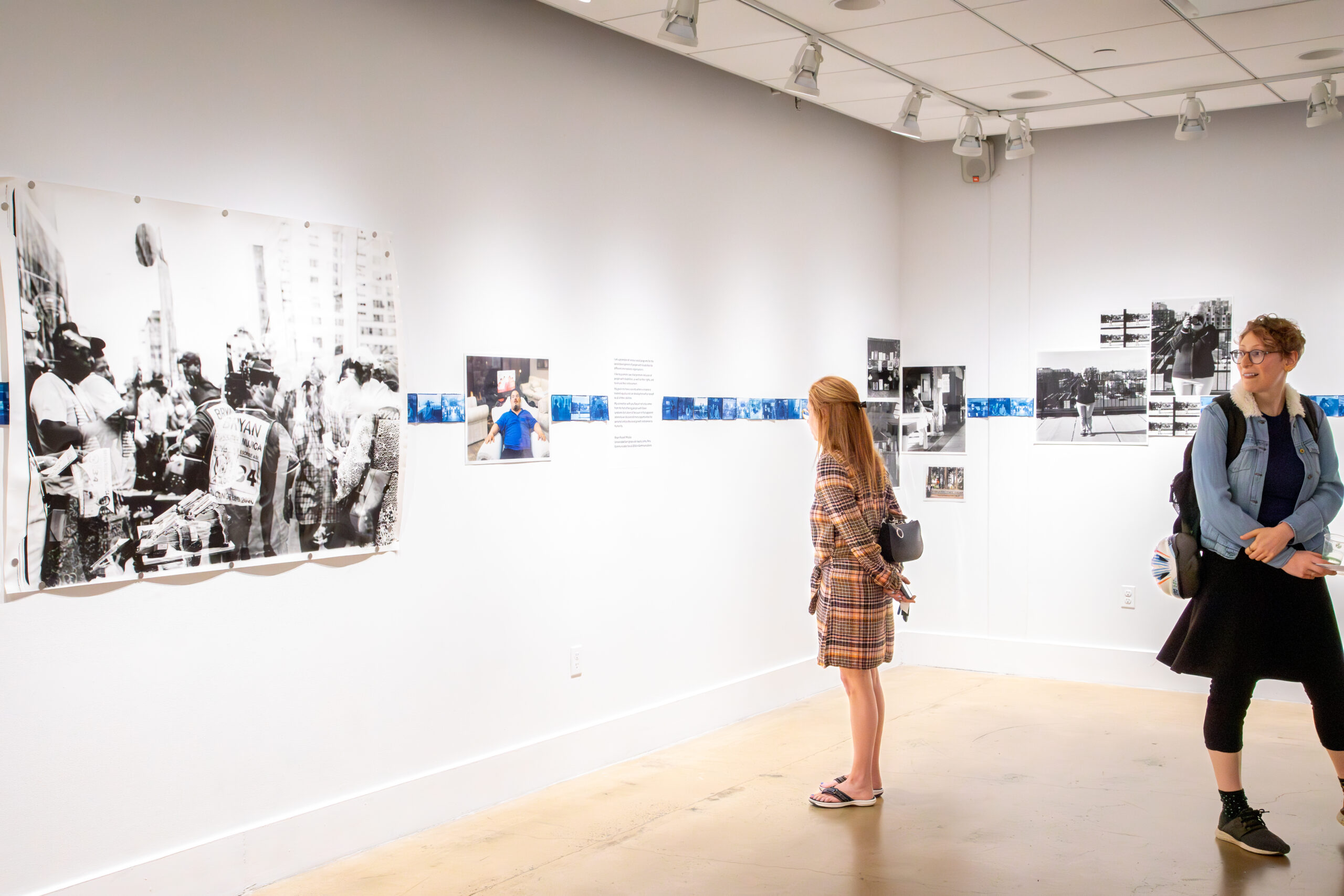 Two individuals stand facing framed artworks displayed on a gallery wall. One of the artworks depicts a black-and-white scene with a group of people. The gallery lighting accentuates the clean and contemporary layout of the space.