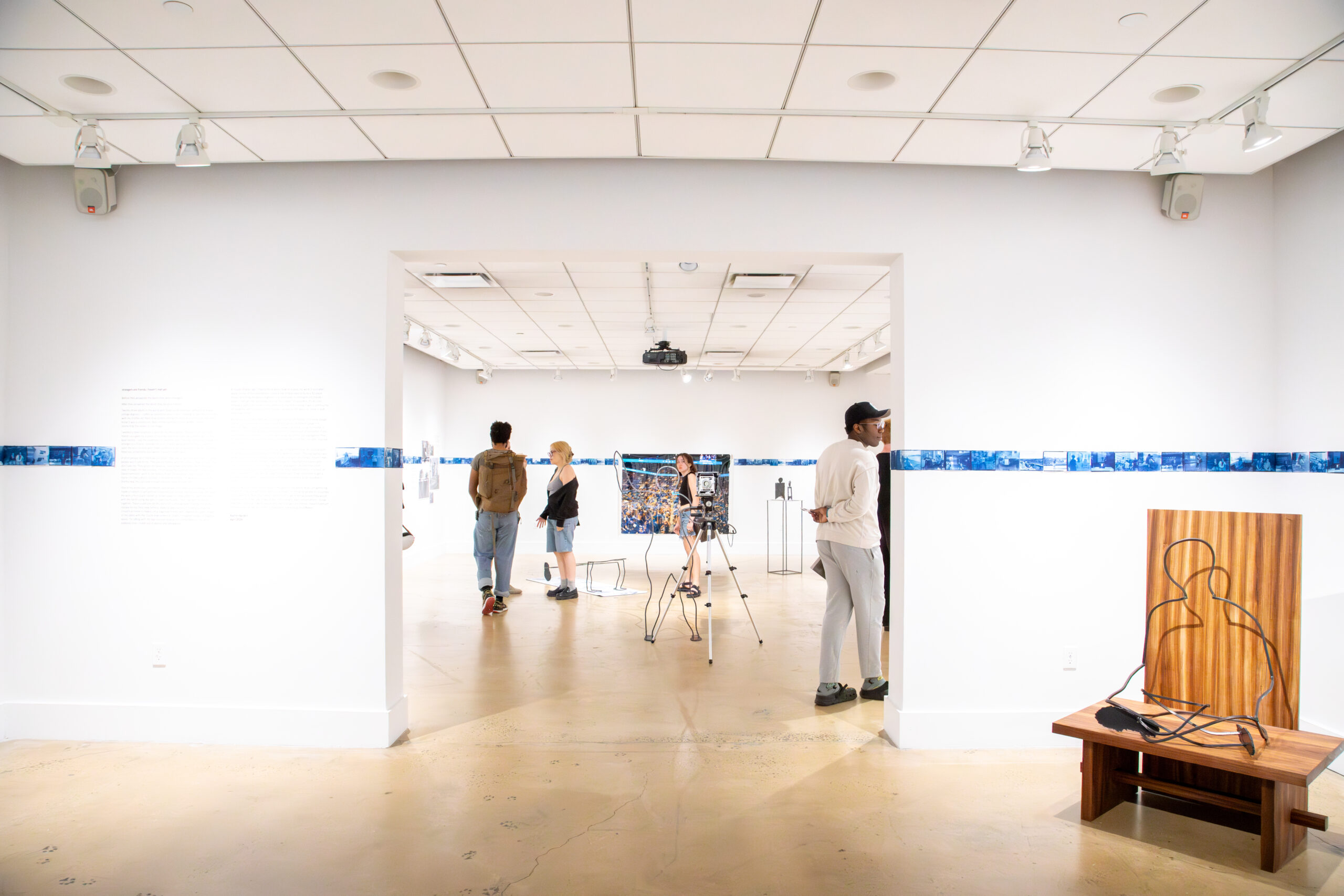 Wide shot of an art gallery showing a variety of people walking and interacting in the space. Framed artworks hang on the white walls, and a small sculpture is visible in the foreground. The room features bright lighting and an open, airy atmosphere.