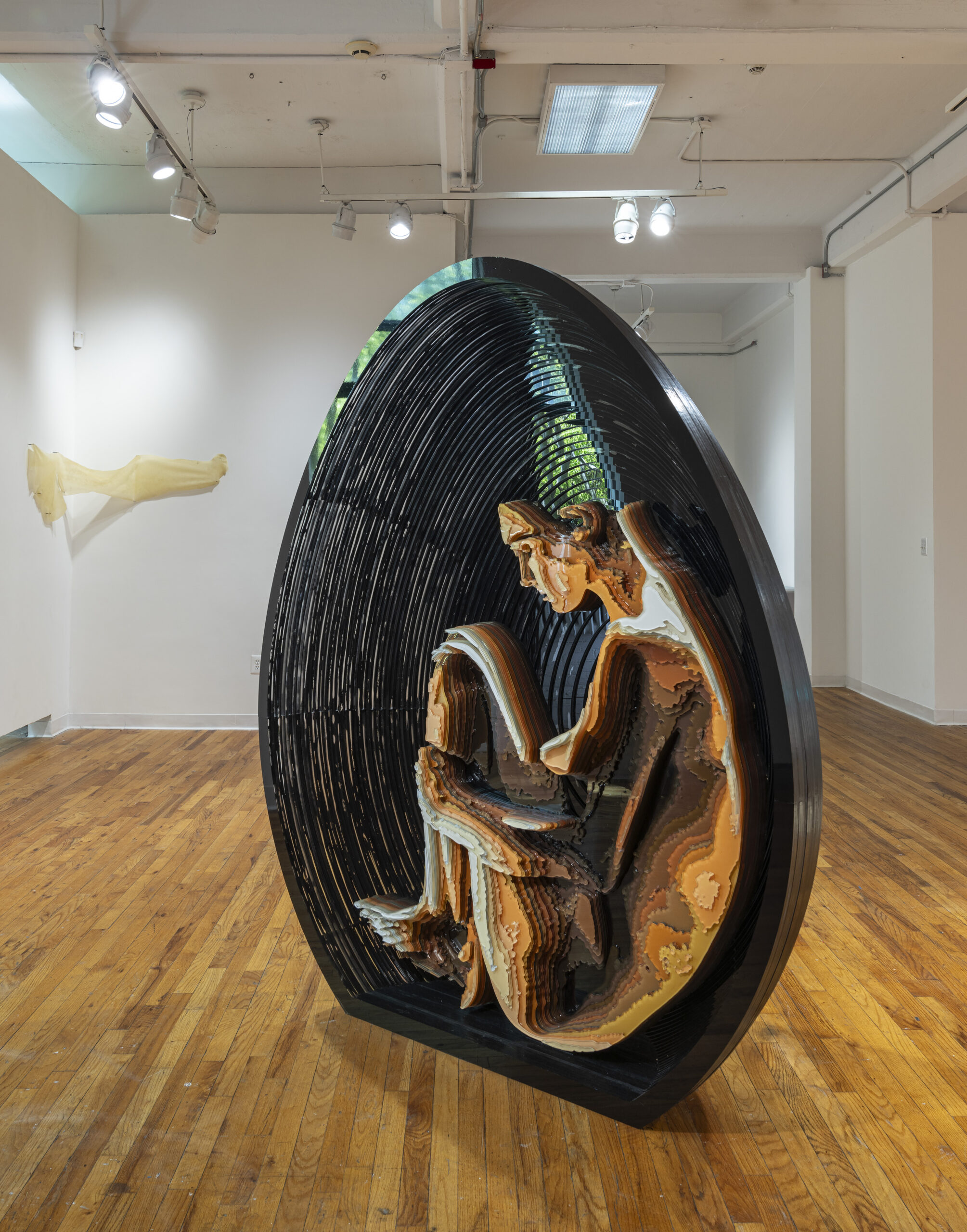 A layered sculptural artwork resembling a seated human figure enclosed within an egg-shaped frame. The sculpture is composed of intricately stacked materials, creating a textured, contour-like effect. The piece sits on a polished wooden floor in a well-lit gallery space with white walls and overhead track lighting.