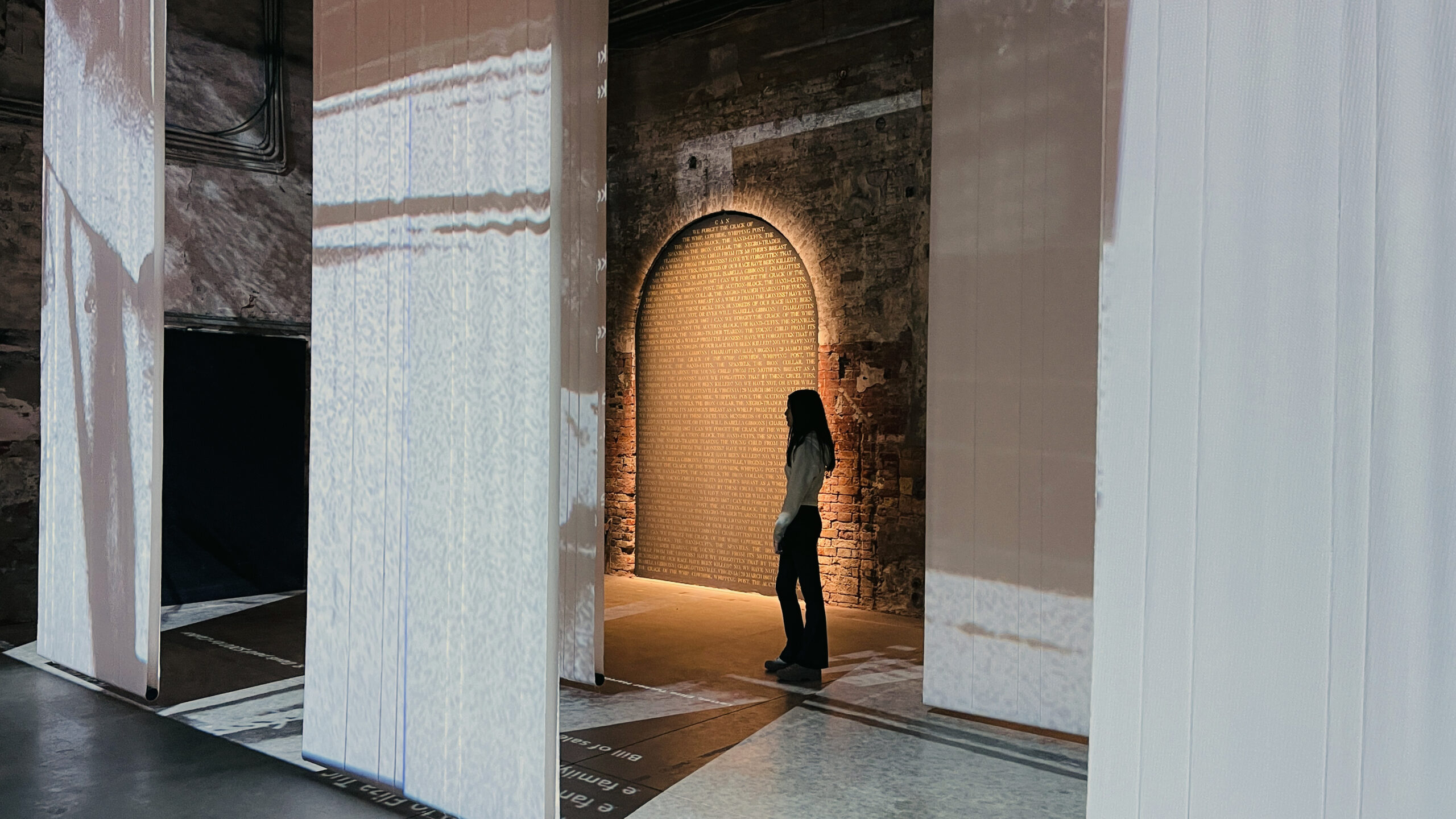 A person silhouetted against a softly illuminated arched wall, inscribed with golden text in a historic brick space. Ethereal projections cascade across hanging white panels and the floor, creating a layered, immersive visual atmosphere. The setting suggests a contemplative installation or exhibition space.