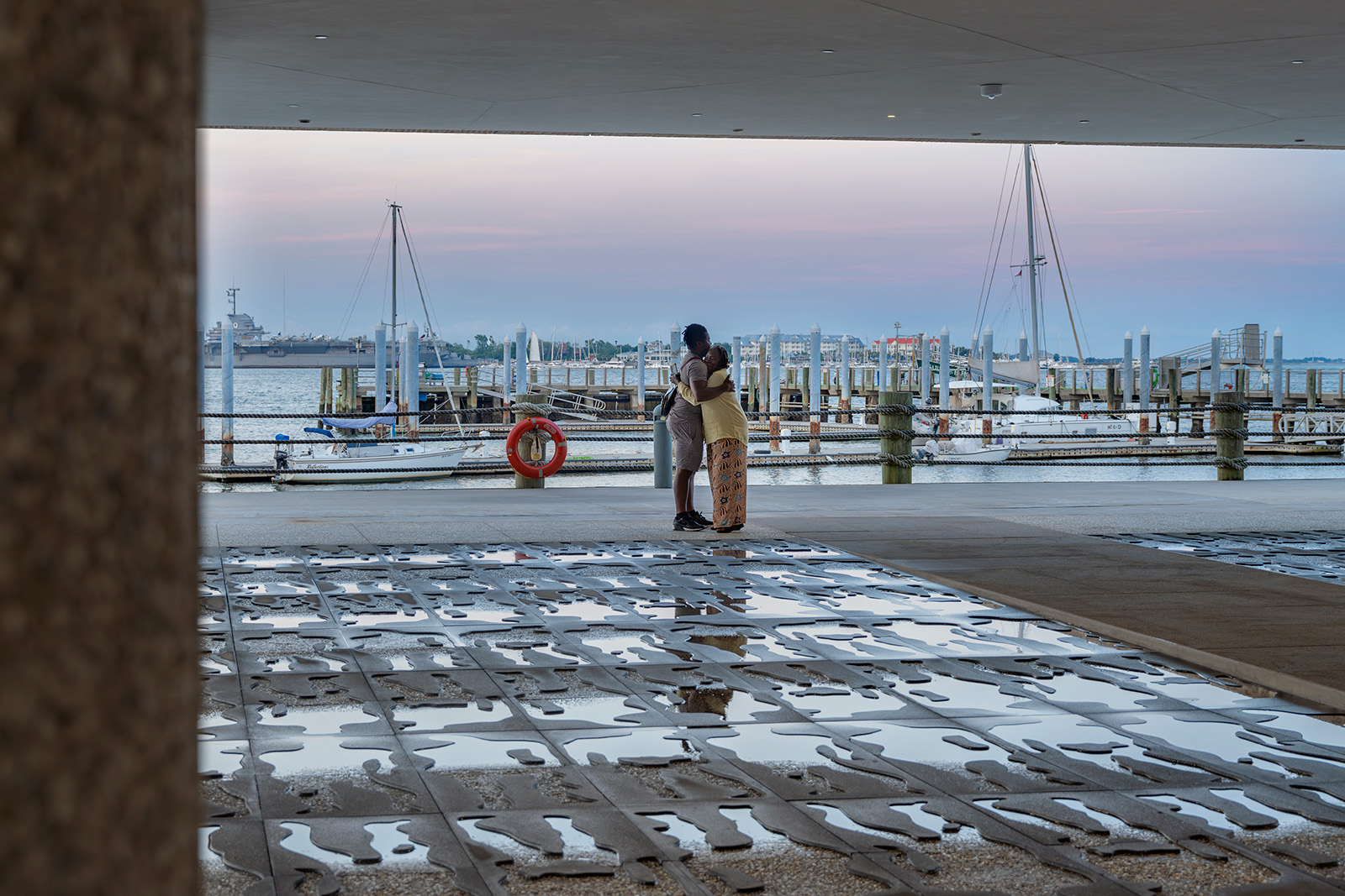 Two people embrace under a modern canopy, framed by a view of a marina with sailboats and a distant ship. The foreground features an artistic, reflective ground surface with abstract cutouts holding small pools of water. The scene is serene, with pastel hues of a sunset over the harbor.