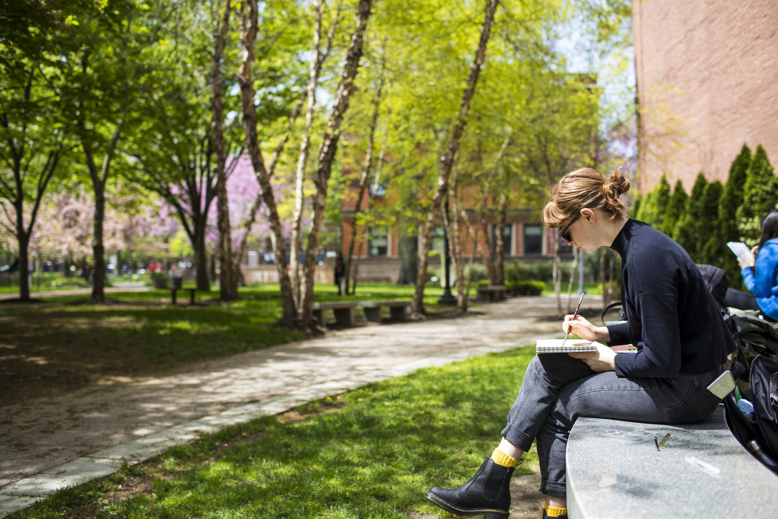 A person sitting outdoors on a stone bench, sketching in a notebook. They are surrounded by a serene and green campus environment, with trees, grassy areas, and pathways in the background. The sunlight filters through the trees, creating a peaceful and reflective atmosphere. The person is focused on their work, suggesting a moment of creativity or study.