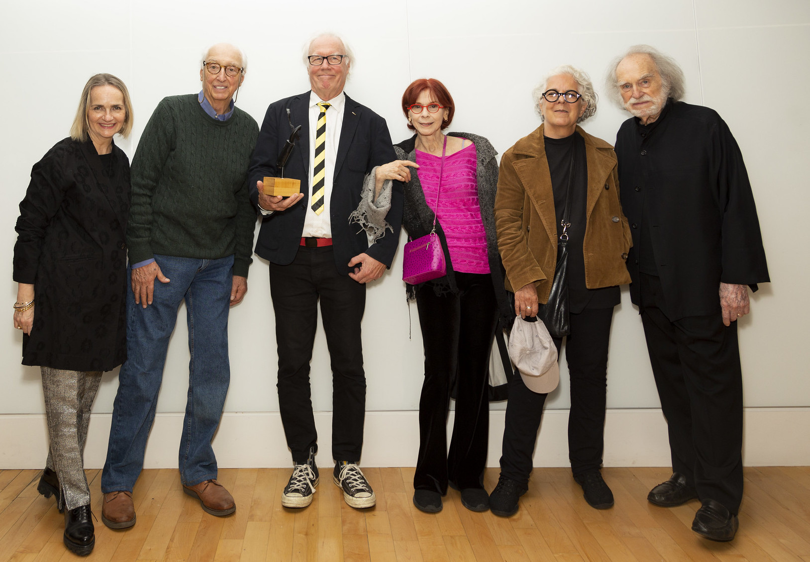 A group photograph featuring six older adults standing in a line, smiling at the camera. The individuals are dressed in a mix of casual and formal attire. The person in the center is holding a small trophy or award. The background is a plain white wall, and the group stands on a light wood floor, creating a warm and celebratory atmosphere.