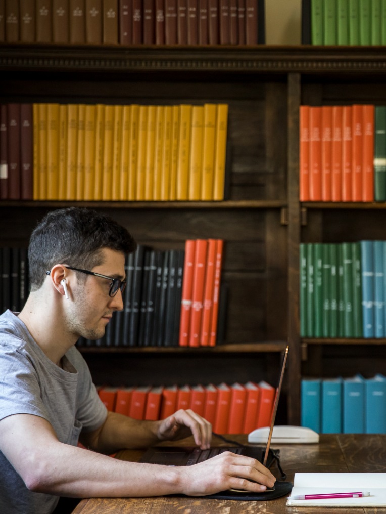 A young man sits at a wooden desk in a library, focused on working on a laptop. He is wearing glasses and AirPods, with short dark hair. Behind him, a large bookshelf is filled with neatly arranged books in various colors, including yellow, red, green, and brown, creating a vibrant backdrop. The scene is quiet and studious, evoking a sense of concentration and academic focus.