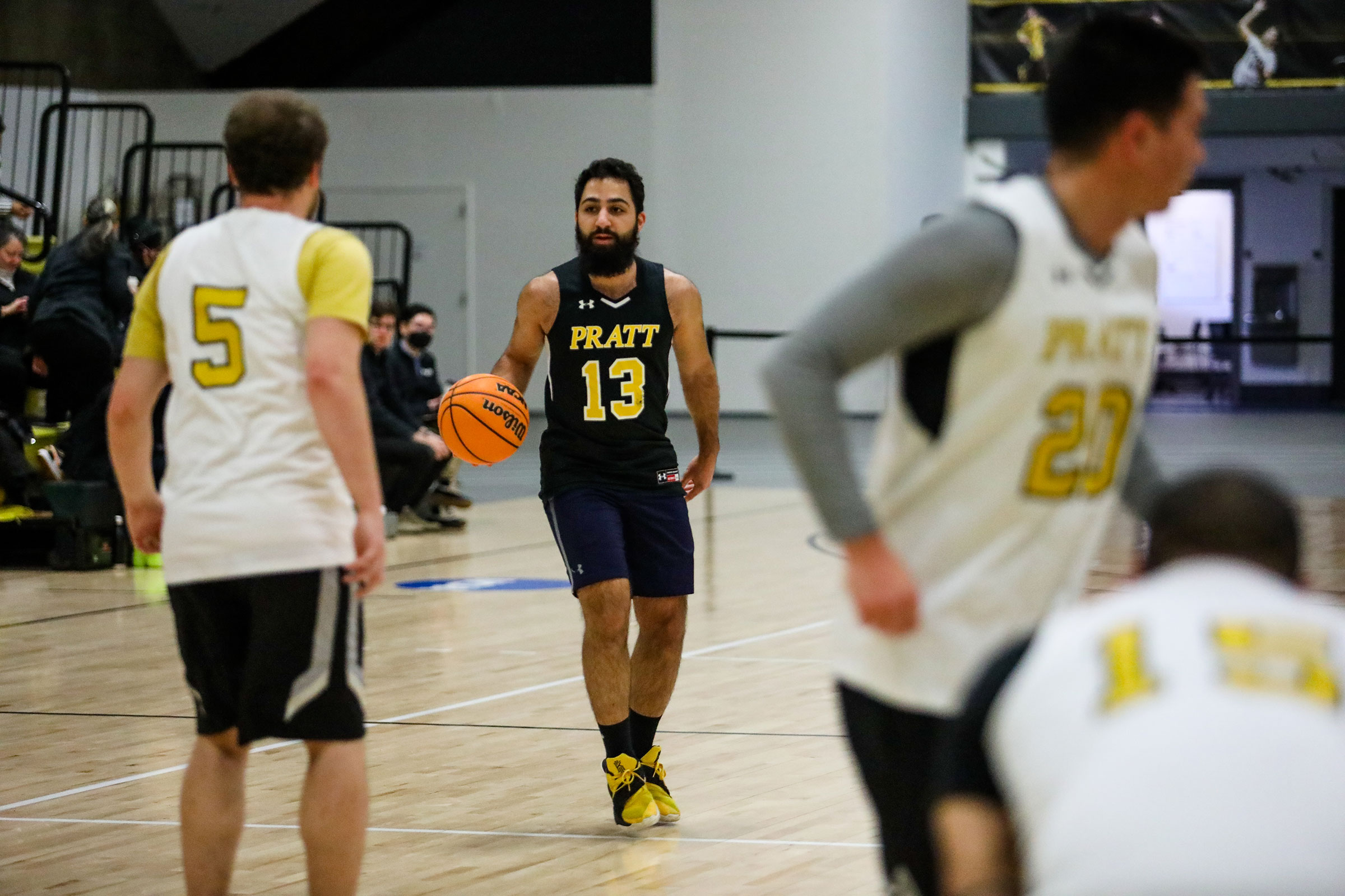 A basketball game in action, featuring a player from Pratt Institute wearing a black jersey with the number 13 in yellow, dribbling the ball down the court. The player has a focused expression and a full beard. Opponents in white jerseys with yellow accents are visible in the foreground, with one player wearing the number 5 facing the Pratt player, and another player wearing number 20 partially visible, moving across the court. The background shows a few spectators sitting in bleachers.