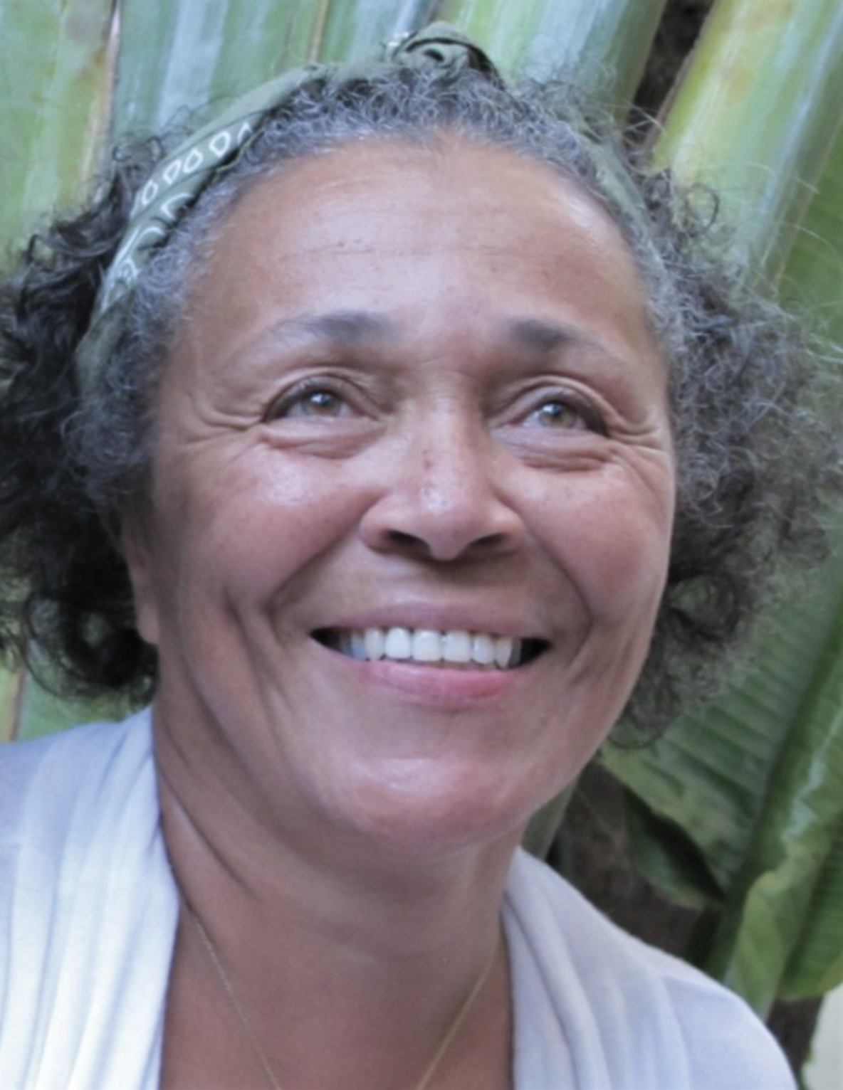 Close-up portrait of Sabine Malebranche, smiling warmly. She has curly hair with a bandana tied around her head, and a green leafy background is visible behind her.