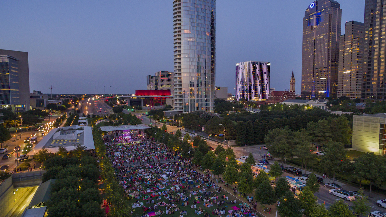 An evening aerial view of Klyde Warren Park in downtown Dallas, with a large crowd gathered on the lawn, enjoying an outdoor event. The cityscape features modern high-rise buildings illuminated by lights, including a vibrant red building and structures with colorful lighting effects. Trees line the park, and cars fill the adjacent streets, contributing to the lively urban atmosphere.