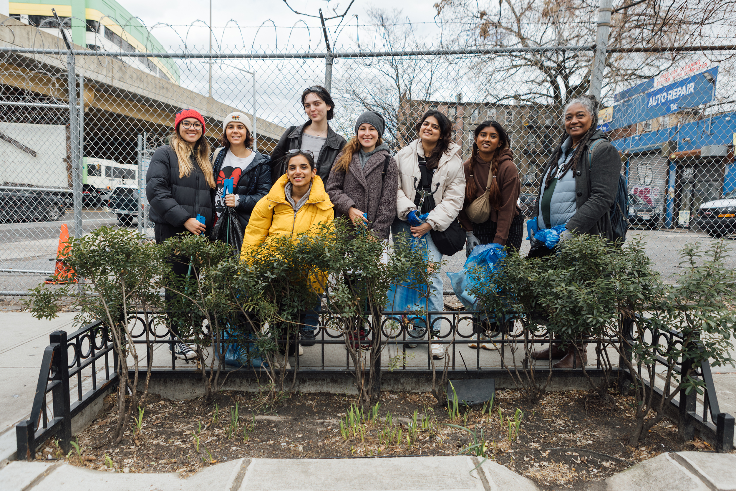 A group of eight individuals standing together and smiling in front of a small, fenced urban green space. They are dressed in winter coats and hats, holding trash bags and litter pickers, with a backdrop of a chain-link fence, an overpass, and an auto repair shop.