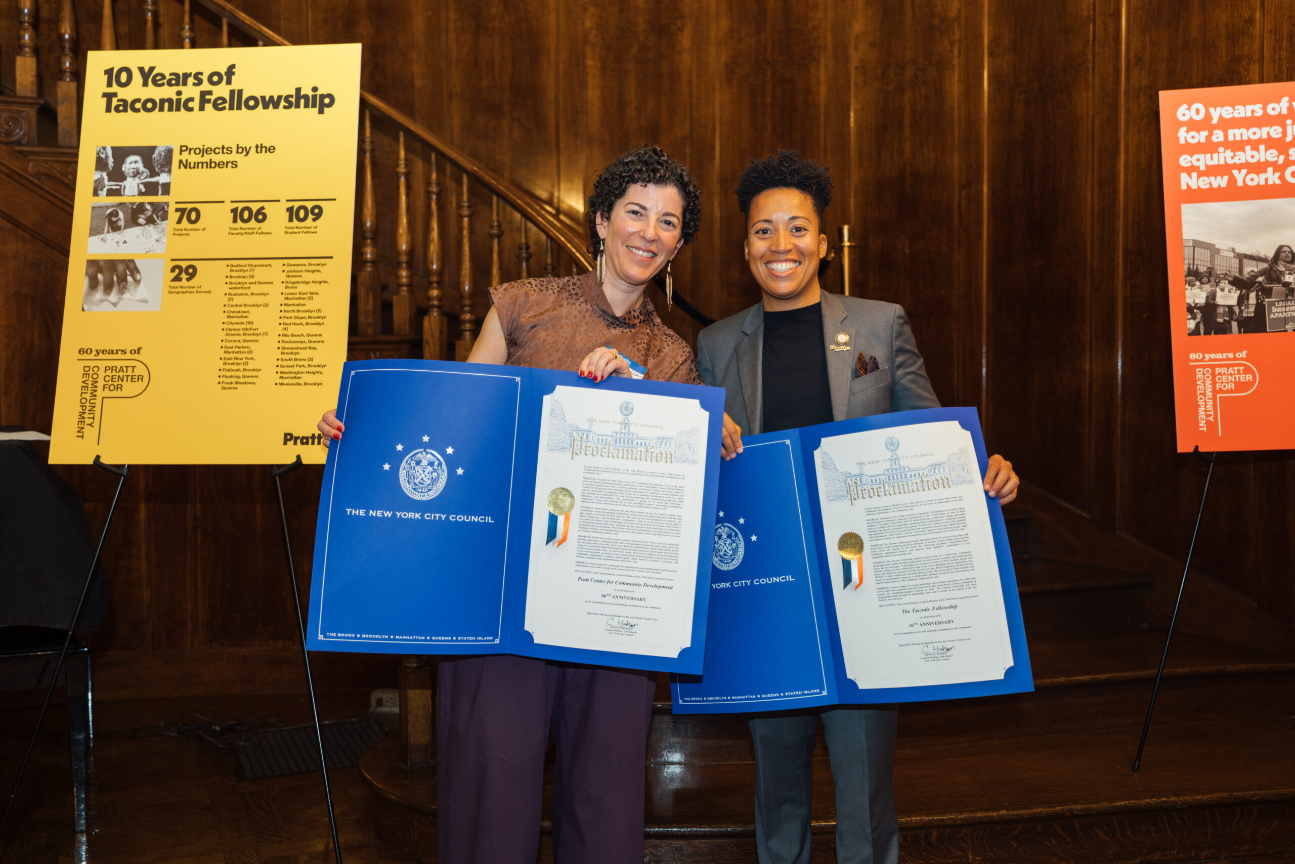 Two people smiling and holding large plaques in a wood-paneled room.