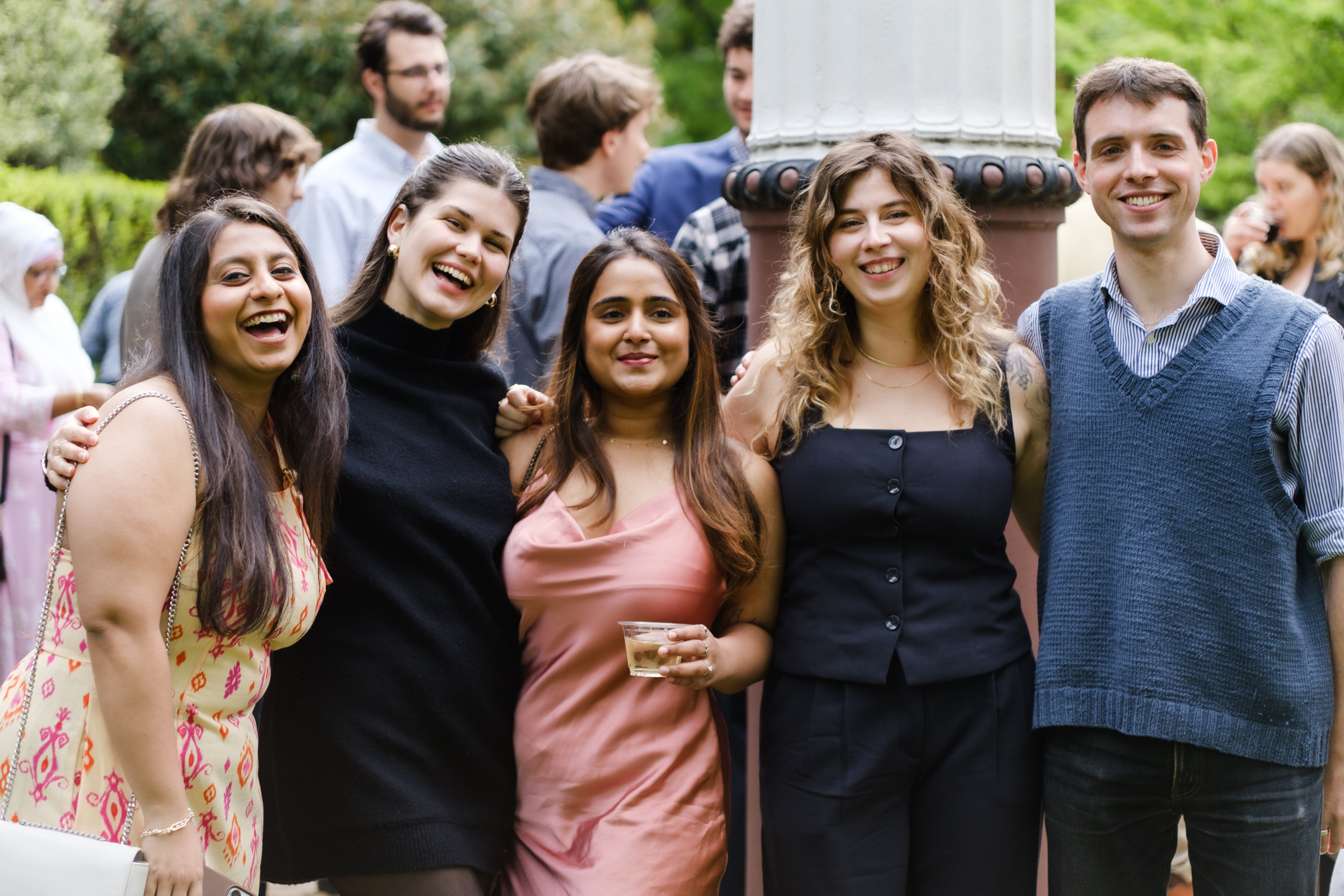 A group of five people standing closely together outdoors, smiling at the camera. The group consists of four women and one man. The woman on the far left is wearing a light-colored dress with pink patterns, the next woman is wearing a black top, the woman in the center is wearing a pink dress and holding a drink, the woman next to her is wearing a black sleeveless top, and the man on the far right is wearing a blue sweater over a striped shirt. They are all standing in front of a large pillar with other people in the background, blurred out.