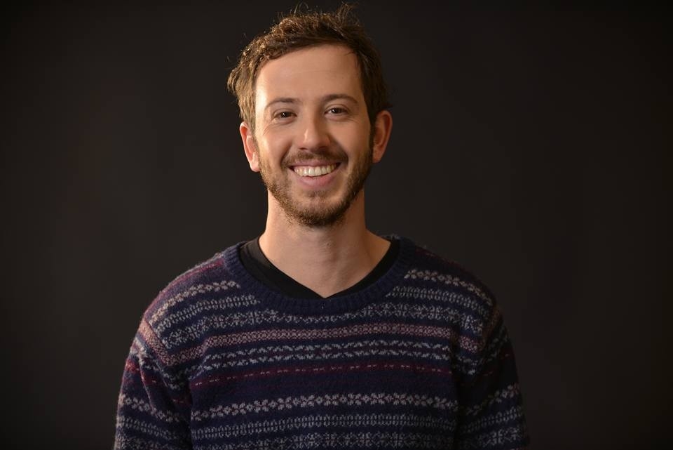 A portrait of a smiling man with short brown hair and a light beard, wearing a dark patterned sweater. He is standing against a dark background.