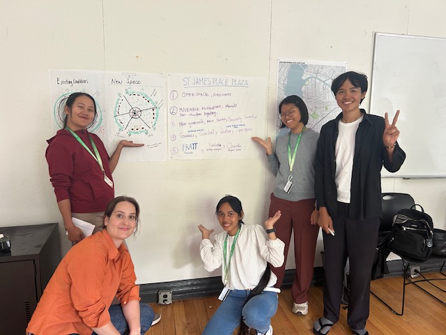 A group of five students posing for a photo in front of their project poster titled "St. James Place Plaza," with one person making a peace sign.