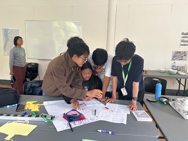 A group of students huddled around a table, working collaboratively on a project with papers and markers spread out.