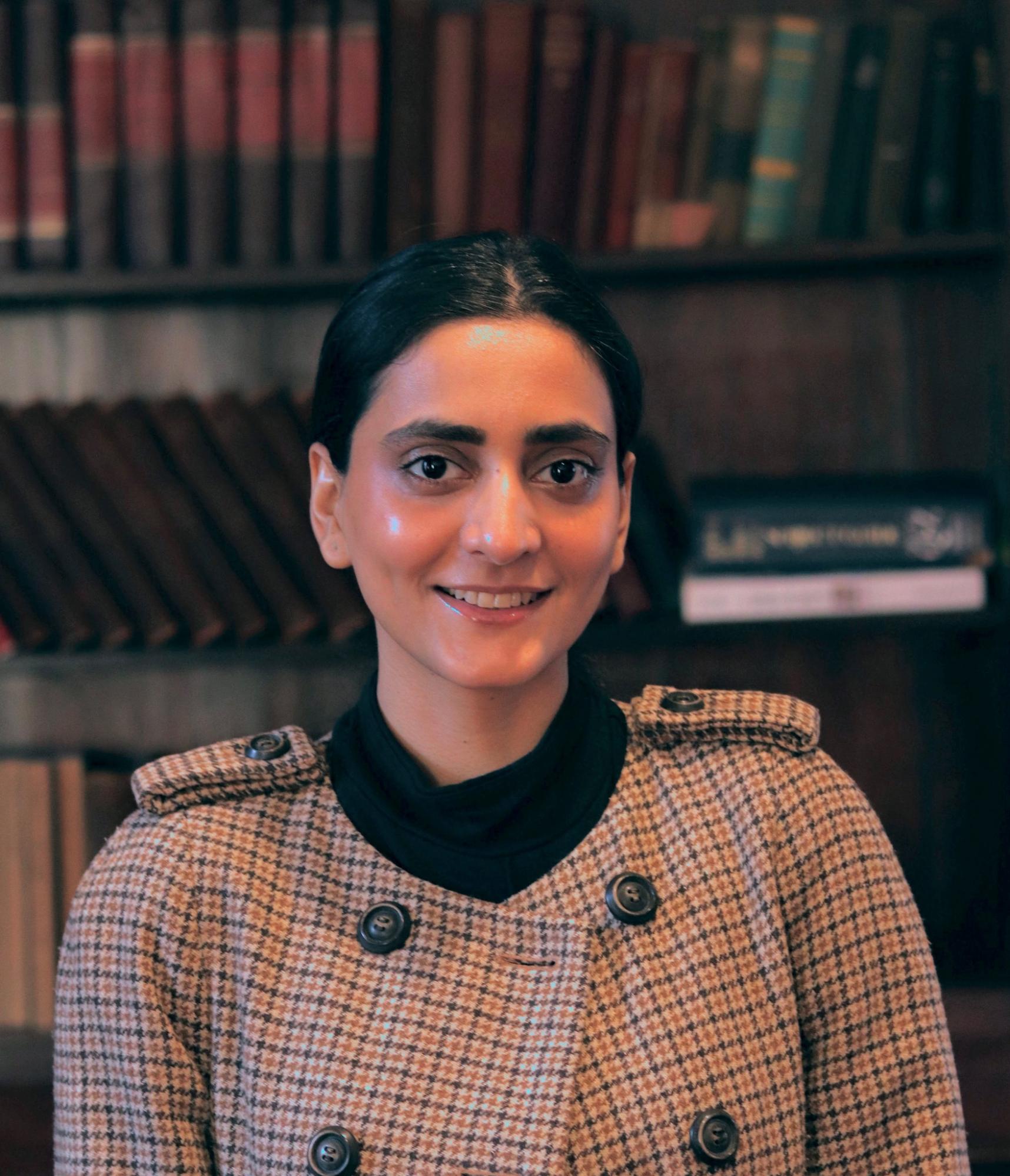 A portrait of a woman standing in front of a bookshelf filled with old hardcover books. She is wearing a brown and beige houndstooth coat with black buttons and a black shirt underneath. The woman is smiling softly and has her dark hair pulled back, with a calm and relaxed expression.