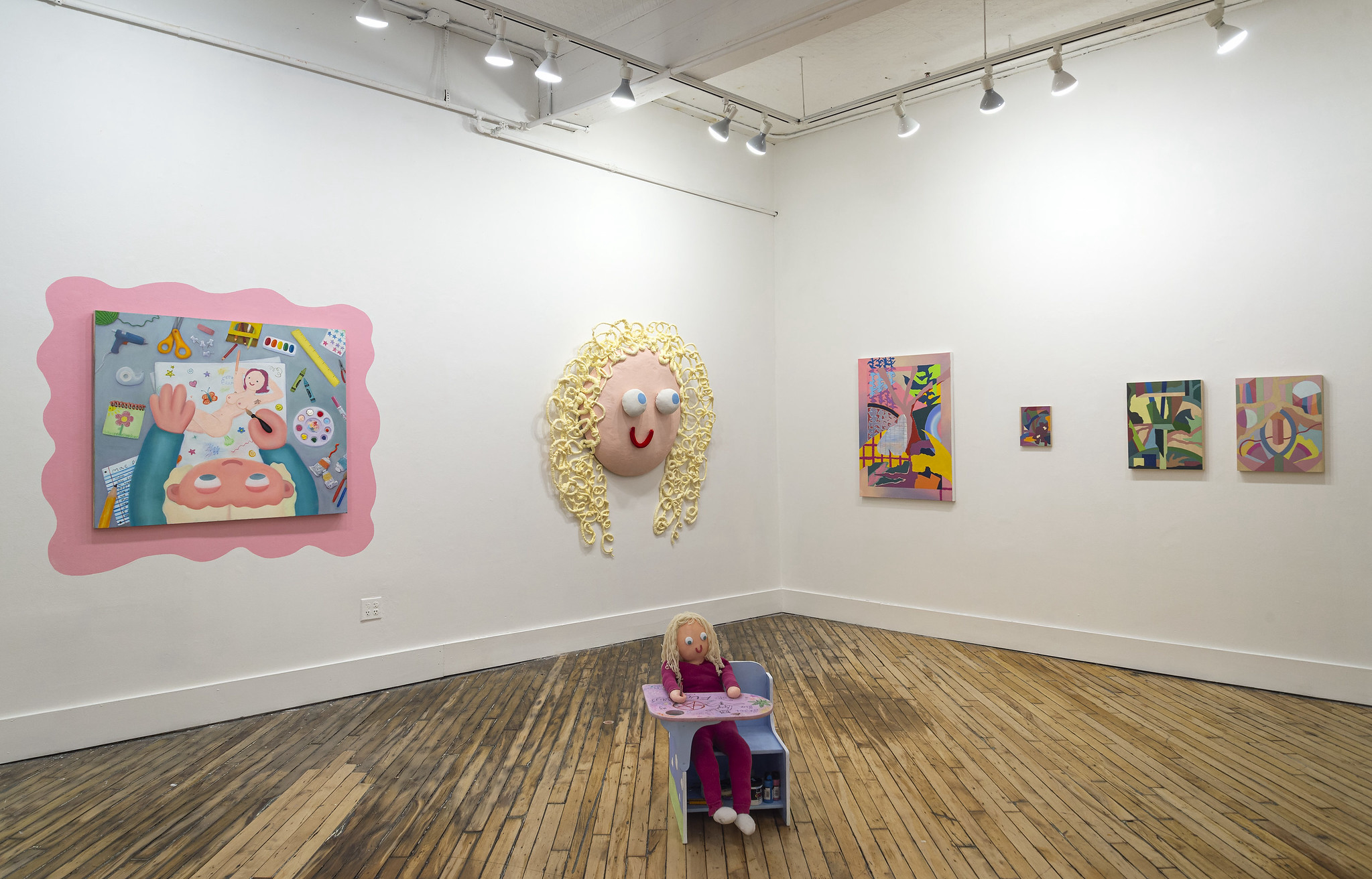 An art gallery corner display featuring a collection of paintings and ceramic sculptures. The walls are adorned with several expressive, colorful paintings, including two large canvases on either side of the corner and several smaller pieces arranged between them. The paintings depict abstract and surreal figures and forms, with vibrant and contrasting colors. In the corner, a white pedestal holds two intricately designed ceramic sculptures, and a ceramic vase with a floral arrangement is placed on the floor nearby. The setting is an intimate gallery space with wooden floors, emphasizing the creative and artistic atmosphere of the exhibition.