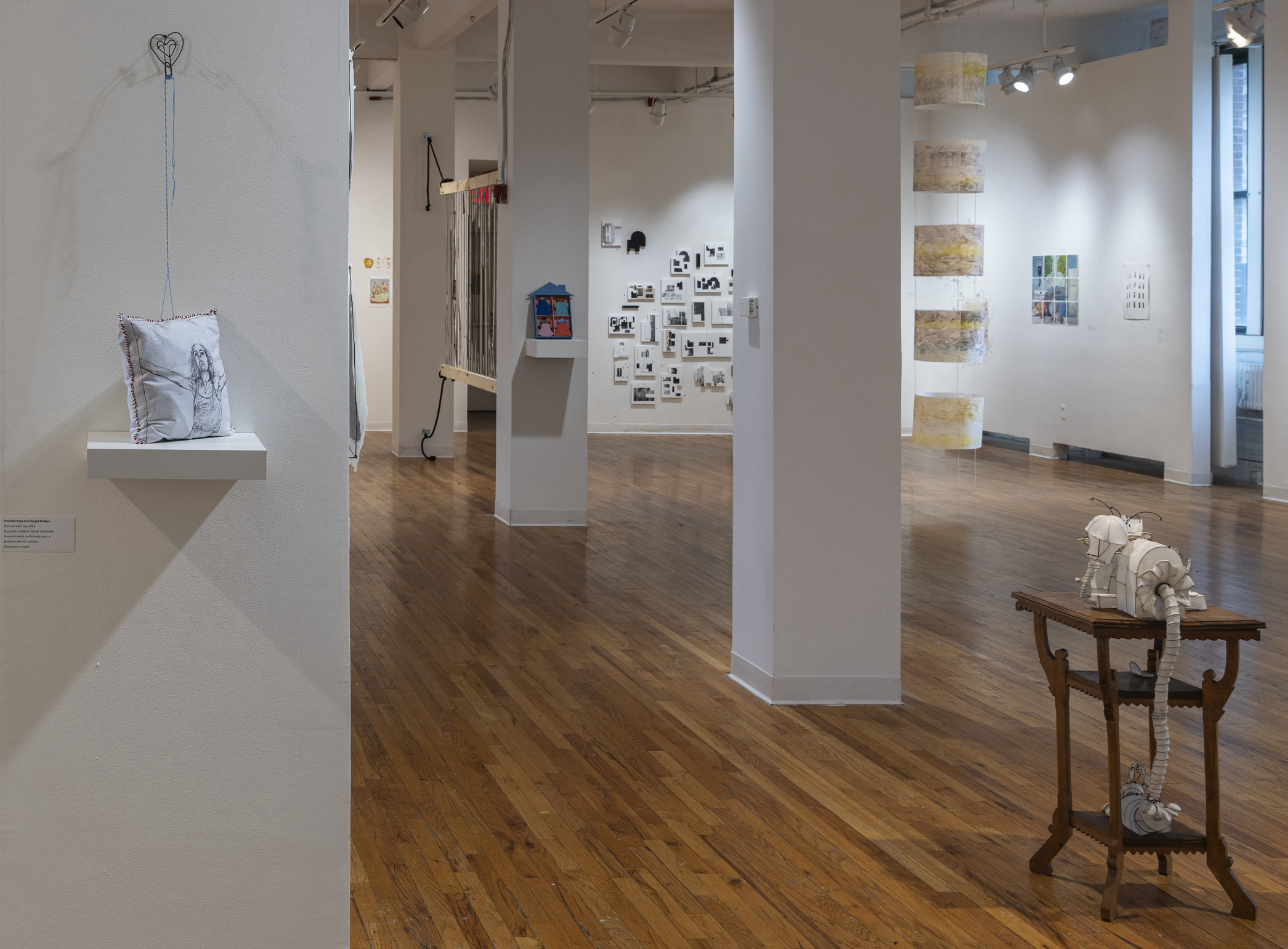 An art gallery space with various installations and artworks. In the foreground, a small white pillow with an embroidered design is displayed on a wall-mounted shelf on the left side. A wooden table on the right side holds a white, sculptural object resembling a segmented, insect-like form. The gallery features wooden floors, with several other artworks and installations visible in the background, including hanging cylindrical prints, wall-mounted drawings, and a variety of multimedia pieces.