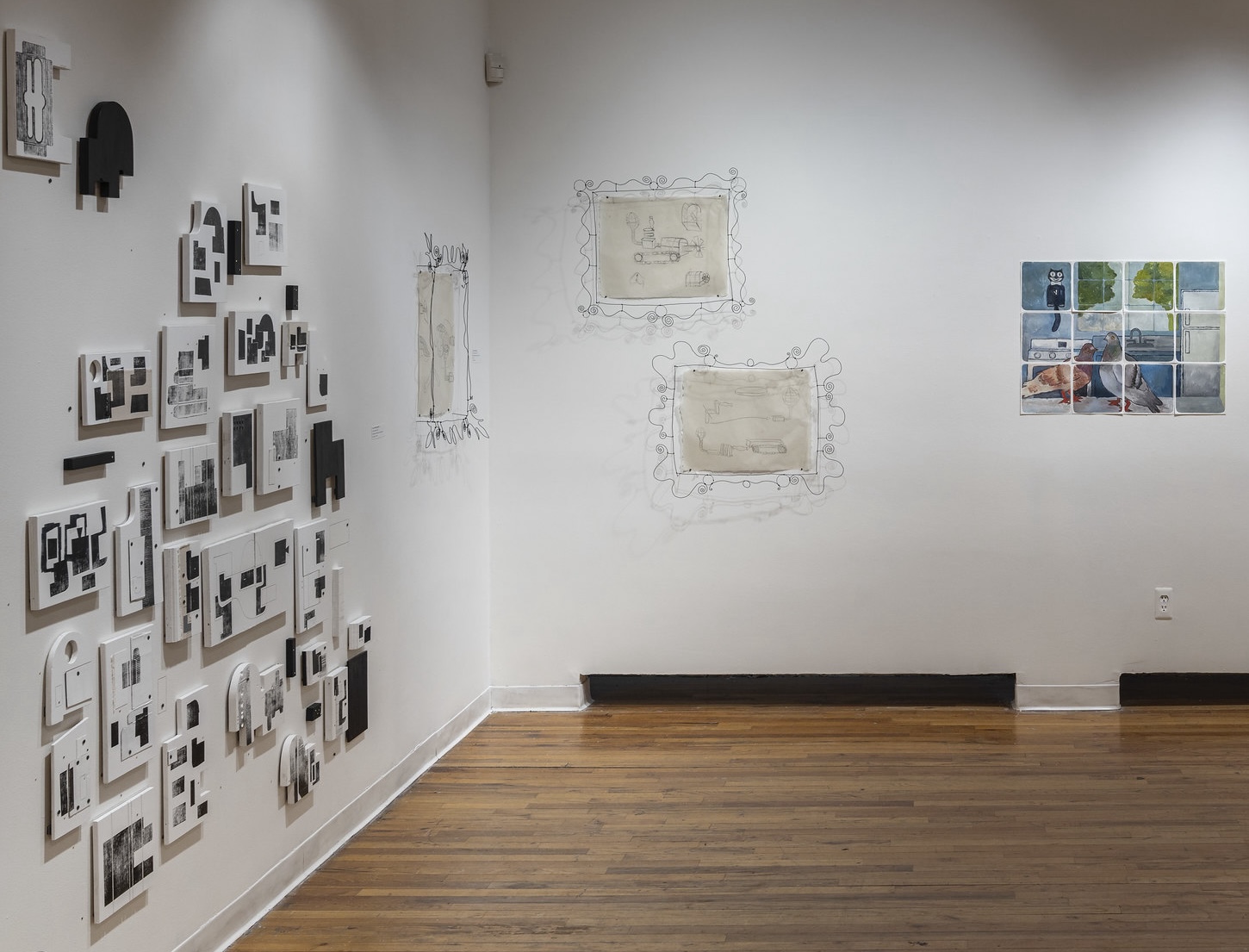 A gallery space with wooden floors and white walls, showcasing a variety of artworks. On the left wall, there is a collection of small, framed black and white abstract prints arranged in a grid-like pattern. To the right, there are two wireframe drawings surrounding framed sketches, giving the impression of ornate, hand-drawn frames. Further to the right, a set of small, square images depicting a pigeon and outdoor scenes are arranged together to form a larger piece. The lighting casts shadows, adding depth to the wireframe drawings.