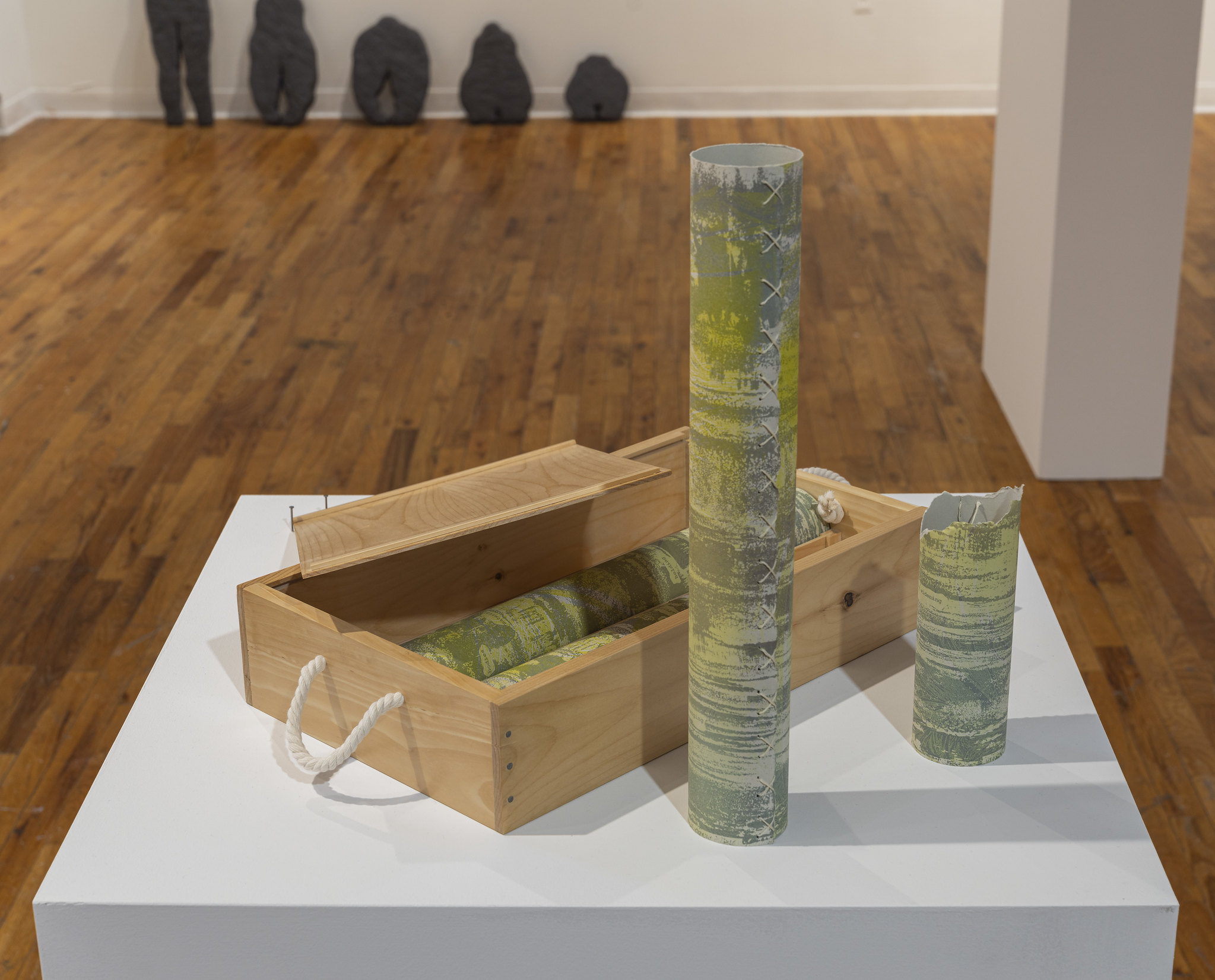 A close-up of a wooden box on a white pedestal, displaying a rolled piece of art with green and yellow hues. Two cylindrical objects, also featuring green and yellow patterns, stand upright beside the box—one is partially broken at the top. The background reveals a wooden floor with several dark, abstract stone-like sculptures lined up against the wall, slightly out of focus.