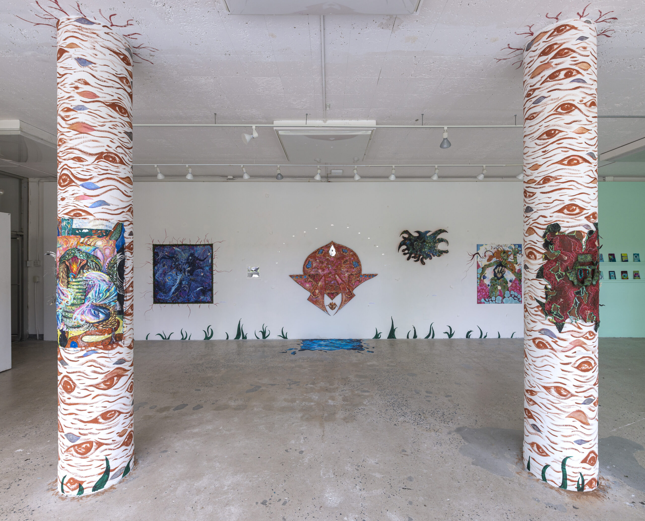 A brightly lit gallery space featuring two large columns in the foreground, intricately wrapped with fabric displaying eye patterns and colorful artwork. The background showcases wall-mounted mixed-media art pieces with organic and abstract shapes, including a large central piece resembling a stylized mask or shield. The gallery floor is concrete, with some painted elements that extend from the wall to the floor.