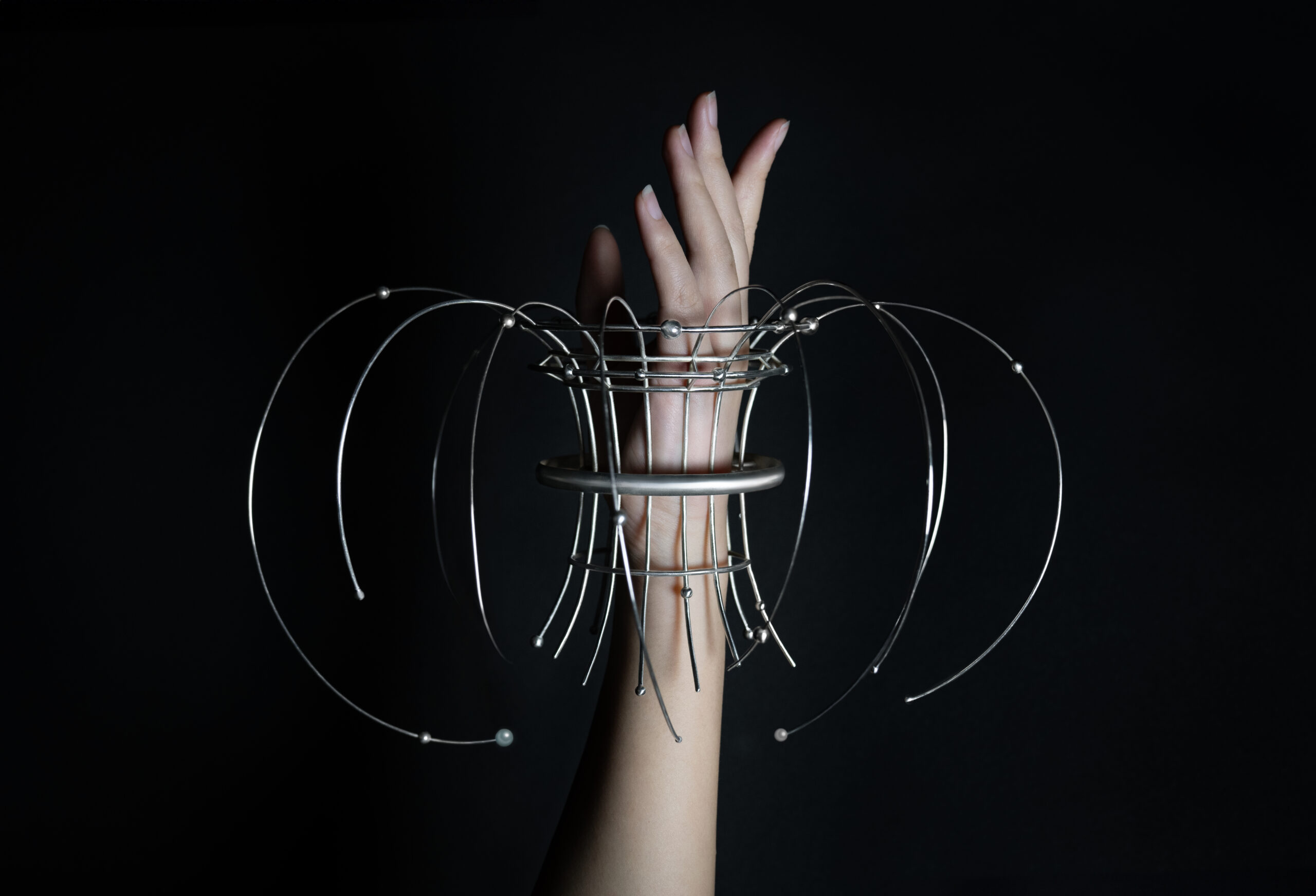 A striking sculptural bracelet made of metal wires and rings, worn on the wrist. The design is avant-garde, featuring circular and curvilinear elements extending outward, creating a dramatic and futuristic effect. The piece is photographed against a dark background, emphasizing the delicate and complex structure.