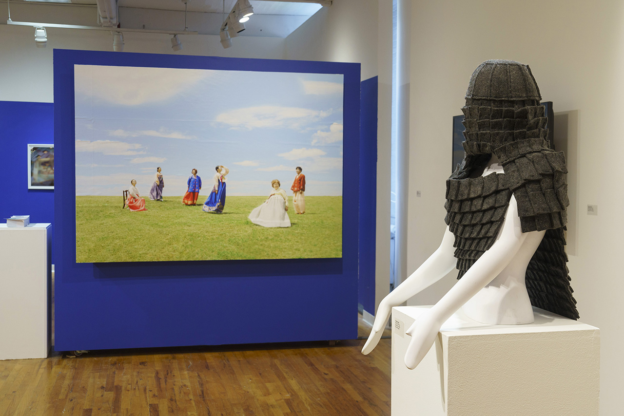 gallery setting, with sculpture in foreground, resembling human torso or bust on a stand, with a head dress, and paintings in the background