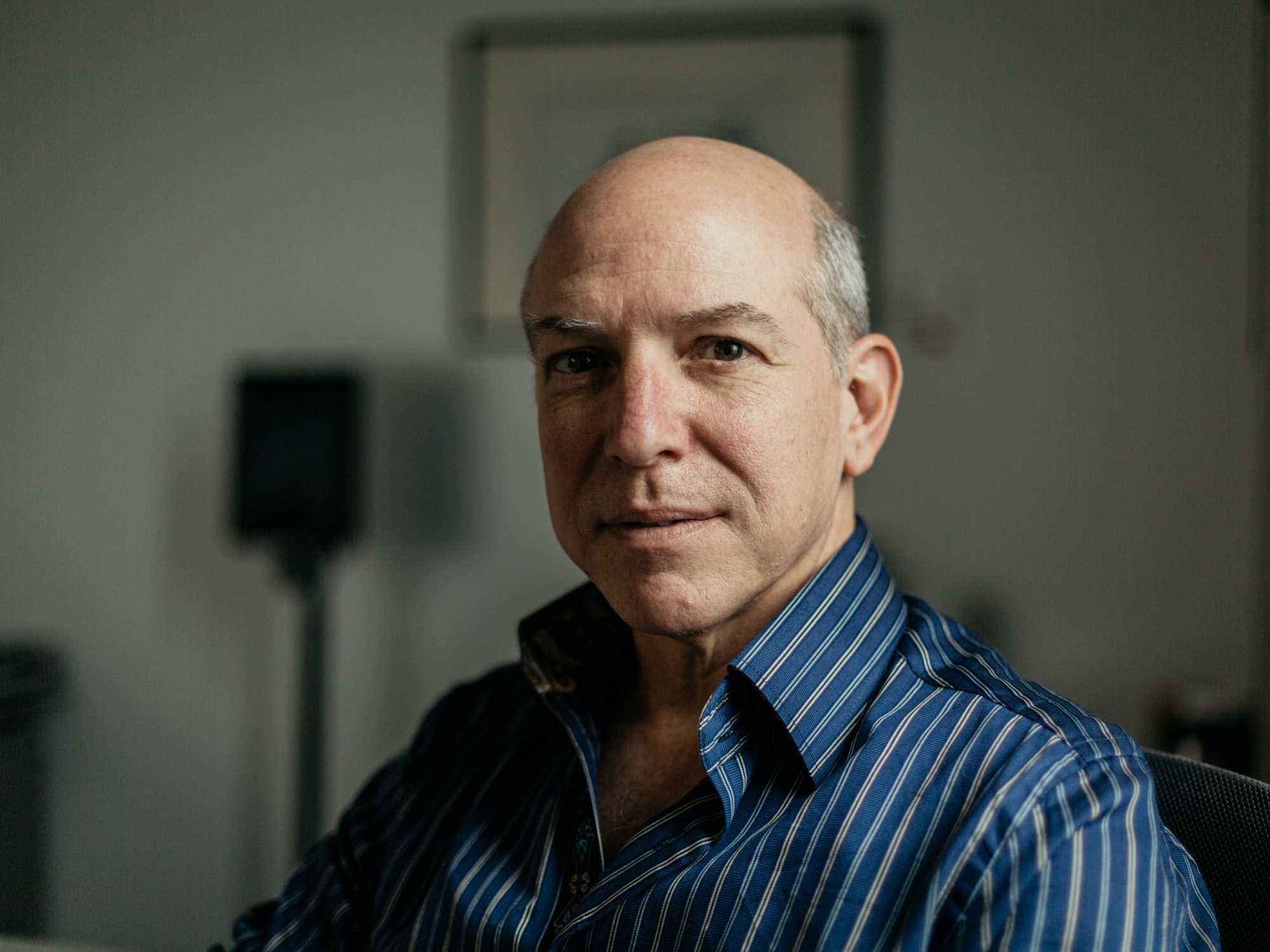 Horizontal portrait of a middle-aged man with a bald head, wearing a striped blue shirt, looking contemplative as he gazes slightly off-camera in an indoor setting.