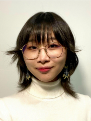 A portrait of a person with short, layered dark hair and round glasses, wearing a light-colored turtleneck. They have star-shaped earrings and a subtle smile, standing in front of a plain background.