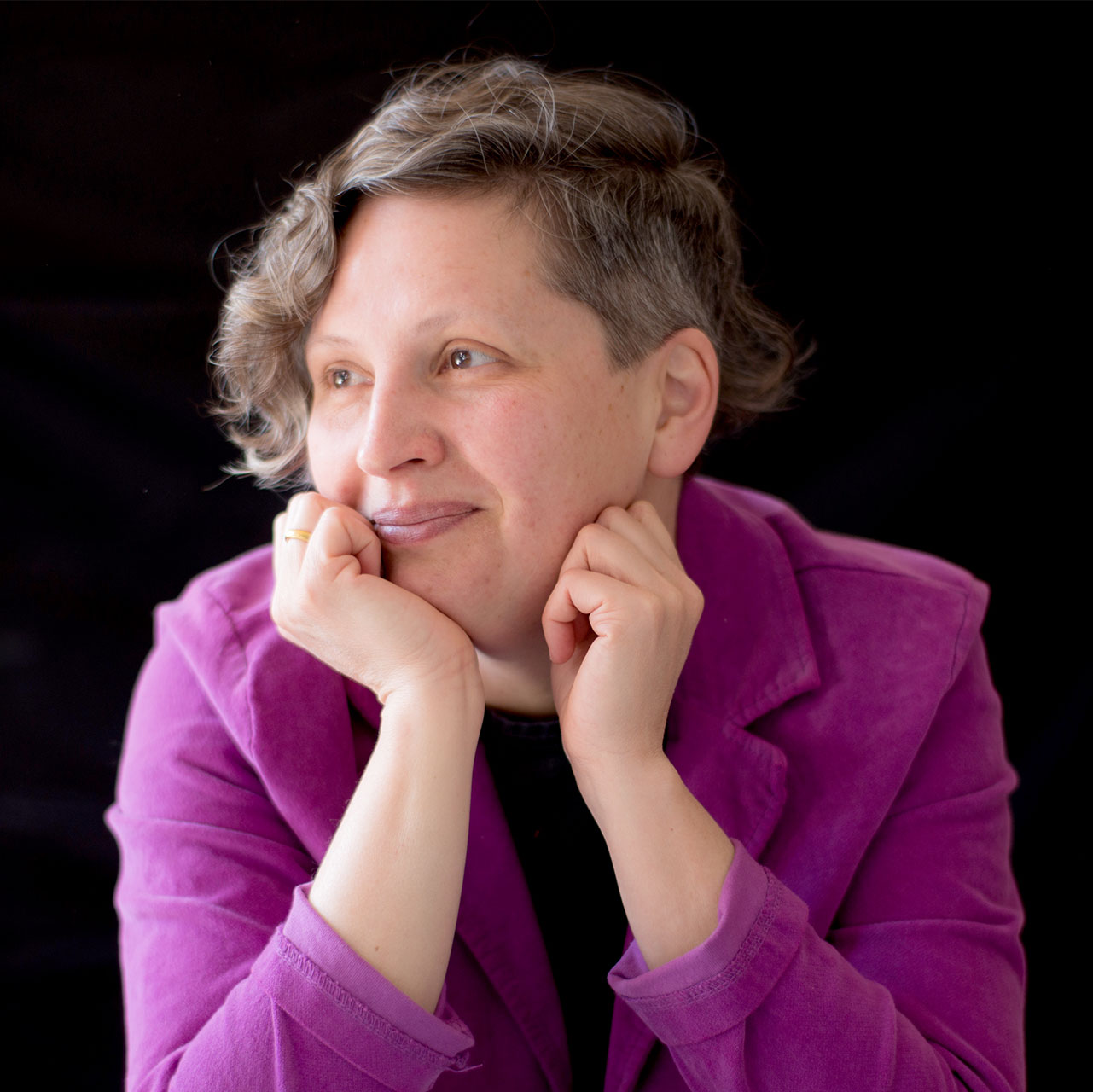 A portrait of a person with short, curly hair wearing a magenta blazer. The individual is gazing off to the side with a gentle smile, resting their chin on their hands. The background is solid black, creating a dramatic and focused composition.