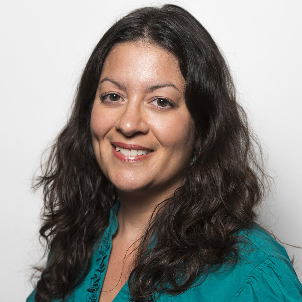 A headshot of a person with long, wavy, dark hair wearing a teal blouse. They are smiling warmly, and the background is plain white, emphasizing the subject's face.