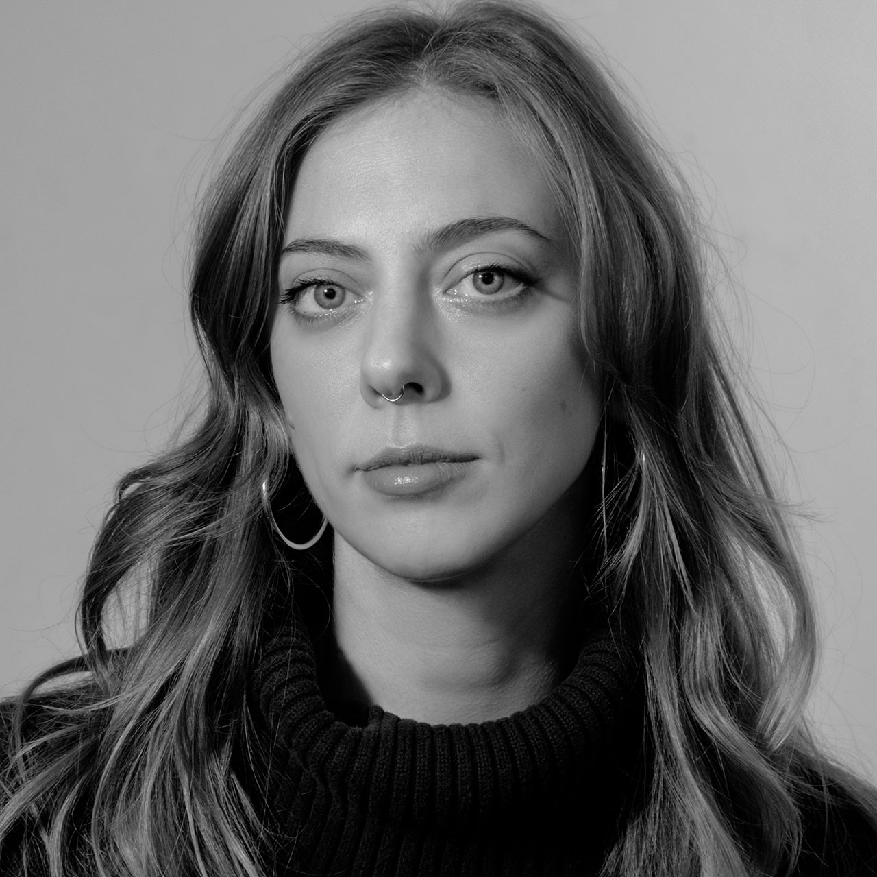 A black-and-white portrait of a person with long, wavy hair, wearing a dark ribbed turtleneck sweater. They have a calm expression, large hoop earrings, and a septum piercing. The background is plain, emphasizing the subject.