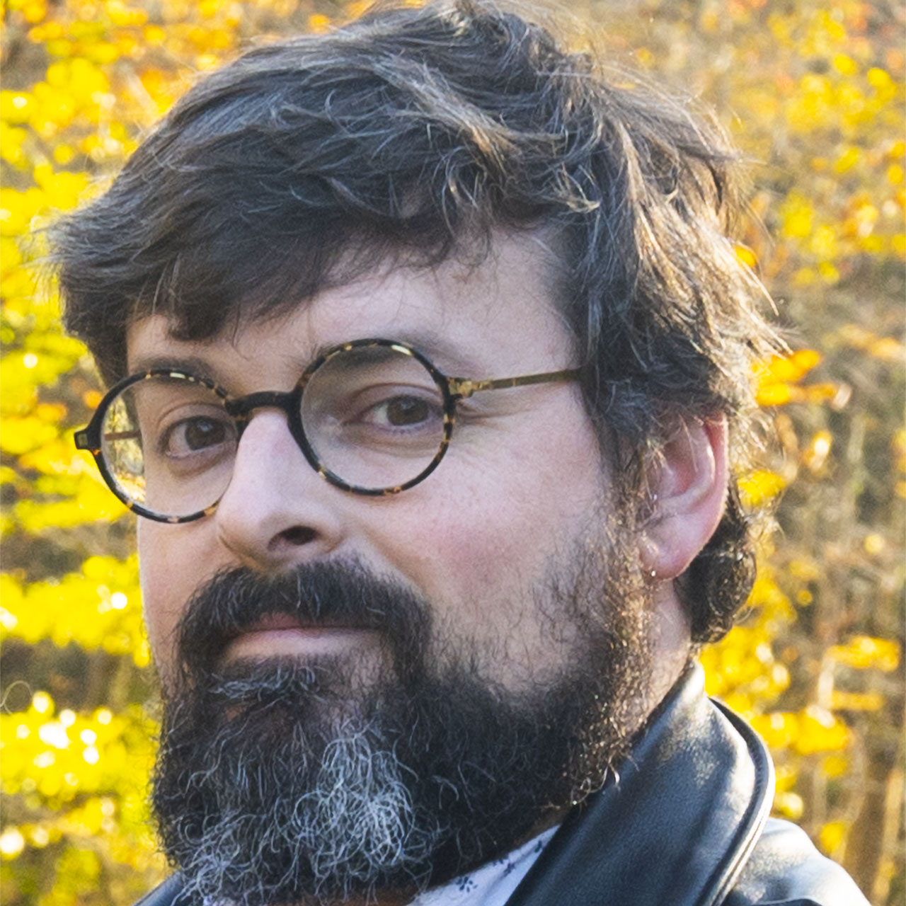 A close-up portrait of a person with short, wavy hair, a full beard, and round tortoiseshell glasses. They are wearing a leather jacket and are outdoors, with a backdrop of vibrant yellow foliage, suggesting a fall setting.