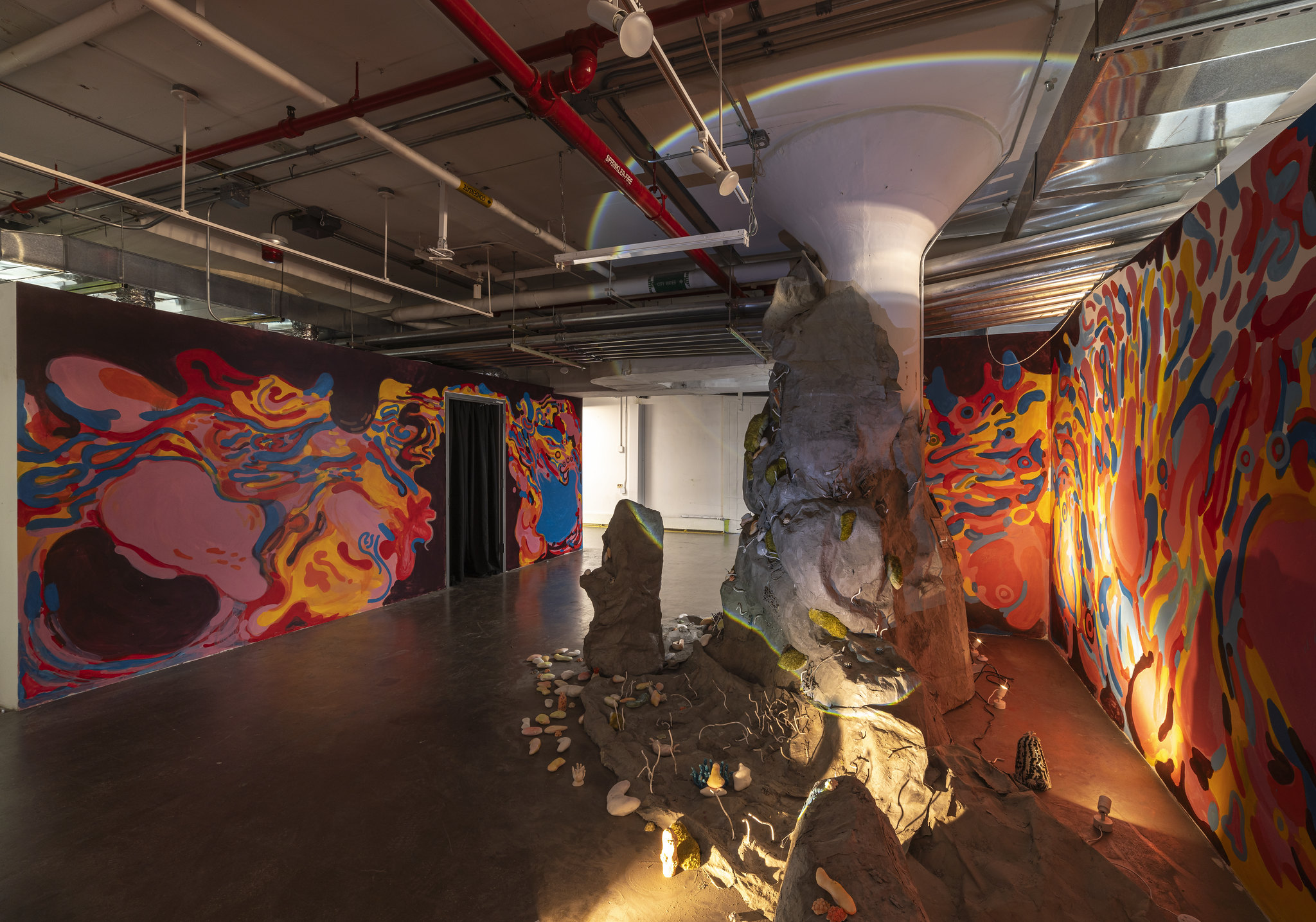 An interior view of an art installation in a dimly lit space. The walls are covered in colorful, abstract murals with swirling patterns in shades of red, orange, blue, and yellow. A large, irregularly shaped sculptural structure, resembling a rocky formation, is positioned in the center of the room, with small objects and elements scattered around its base, creating a surreal and immersive environment. The ceiling features exposed pipes and industrial lighting, adding to the installation's atmospheric setting.