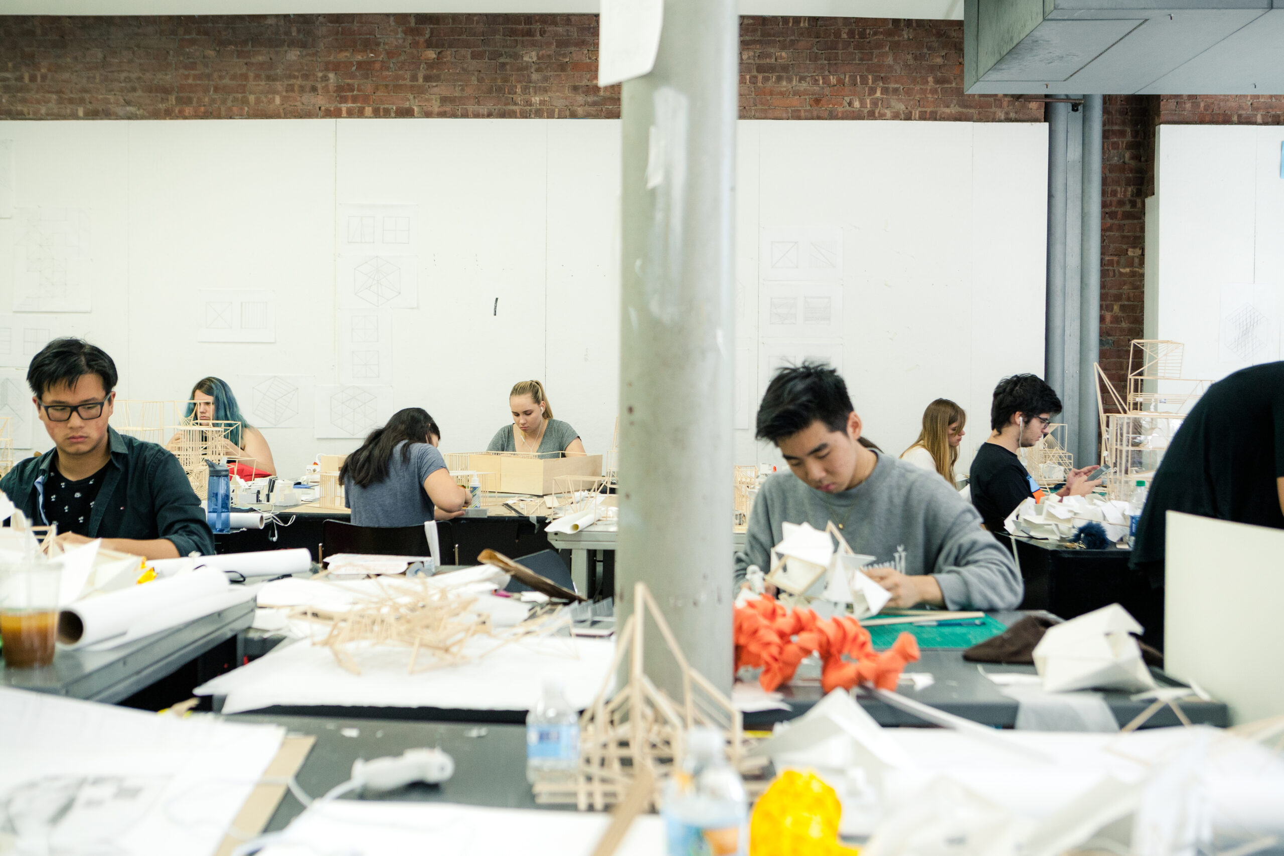 A classroom scene featuring architecture or design students working on scale models at their desks. The room is filled with materials such as wood, paper, and crafting tools, with sketches and diagrams pinned on the walls. The atmosphere is focused and creative, with students deeply engaged in their projects.