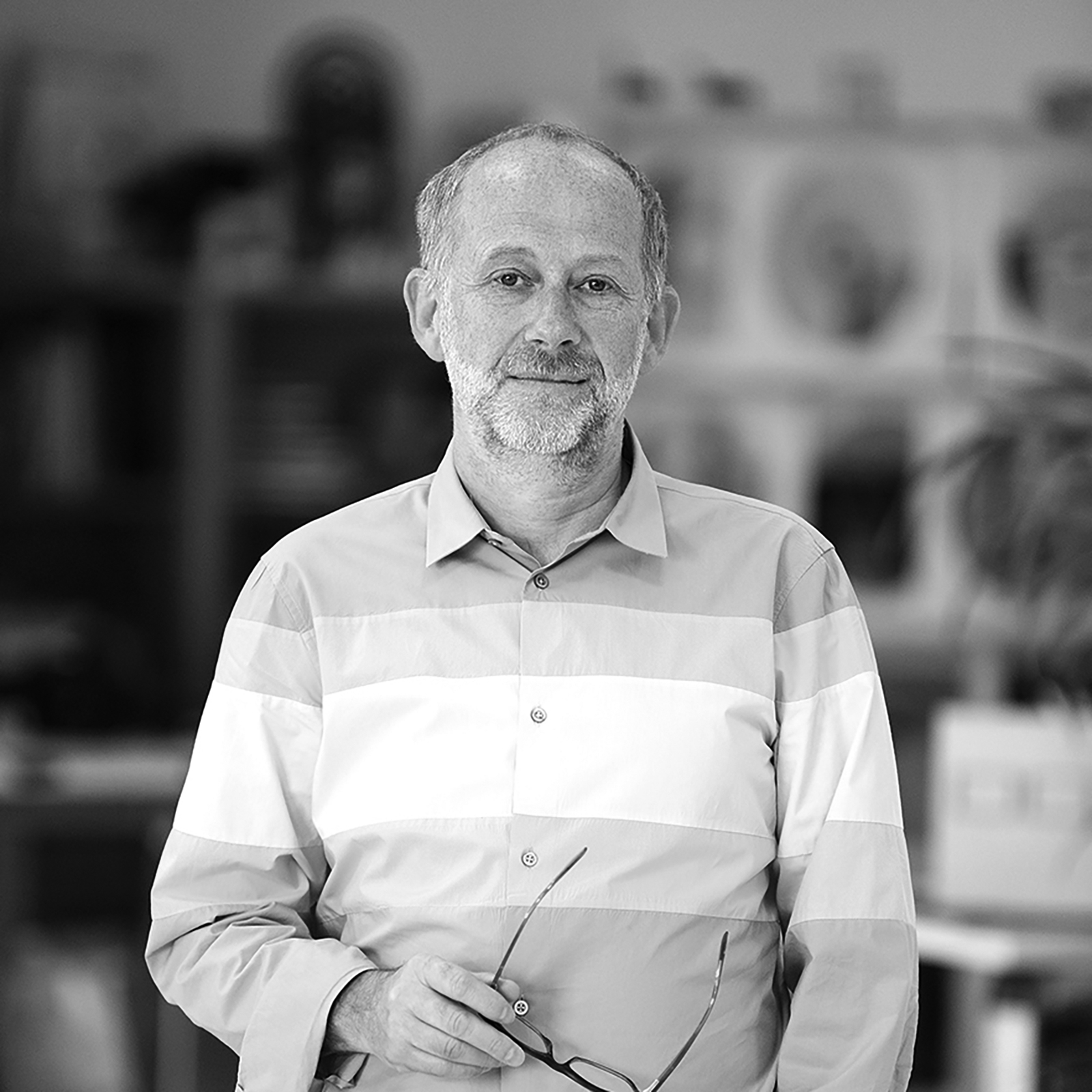 A black-and-white portrait of Constantin Boym, standing in an indoor setting, wearing a striped button-up shirt, holding his glasses.