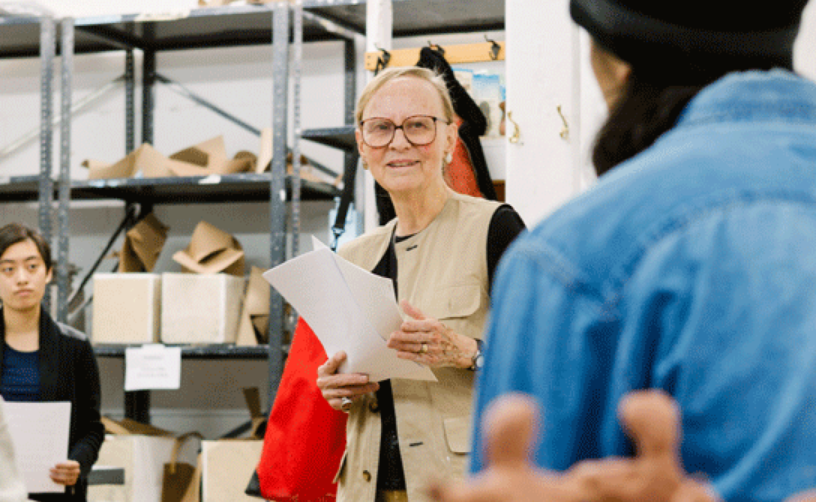 An instructor with short, light-colored hair and glasses stands in a classroom, holding a sheet of paper and addressing students. Behind them, shelves with various materials, including cardboard and paper boxes, are visible. Another person listens attentively to the instructor's speech in the foreground, dressed in a blue denim shirt.