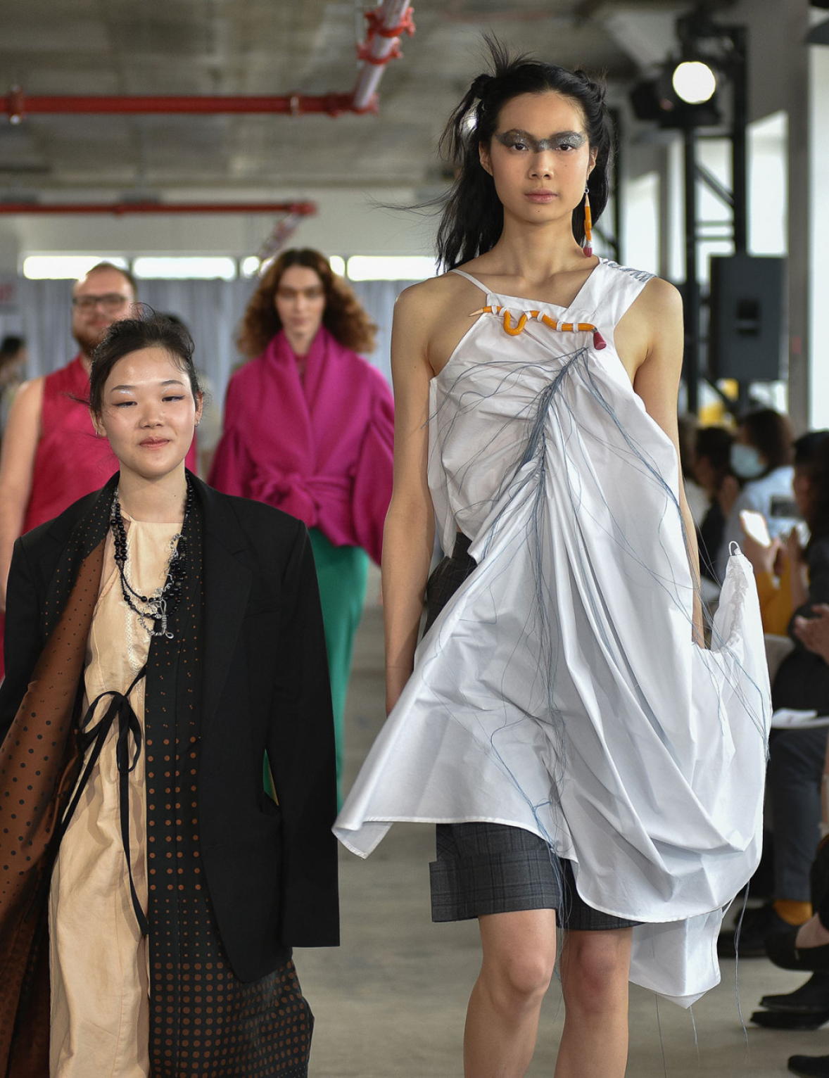 Fashion runway scene with models walking forward. The front model on the right wears a white, asymmetrical dress with flowing blue thread details and a statement orange earring, while the person beside them is dressed in a layered outfit with a black blazer and a beige dress with polka dot patterns. Two other models are visible in the background, one wearing a vibrant magenta wrap.