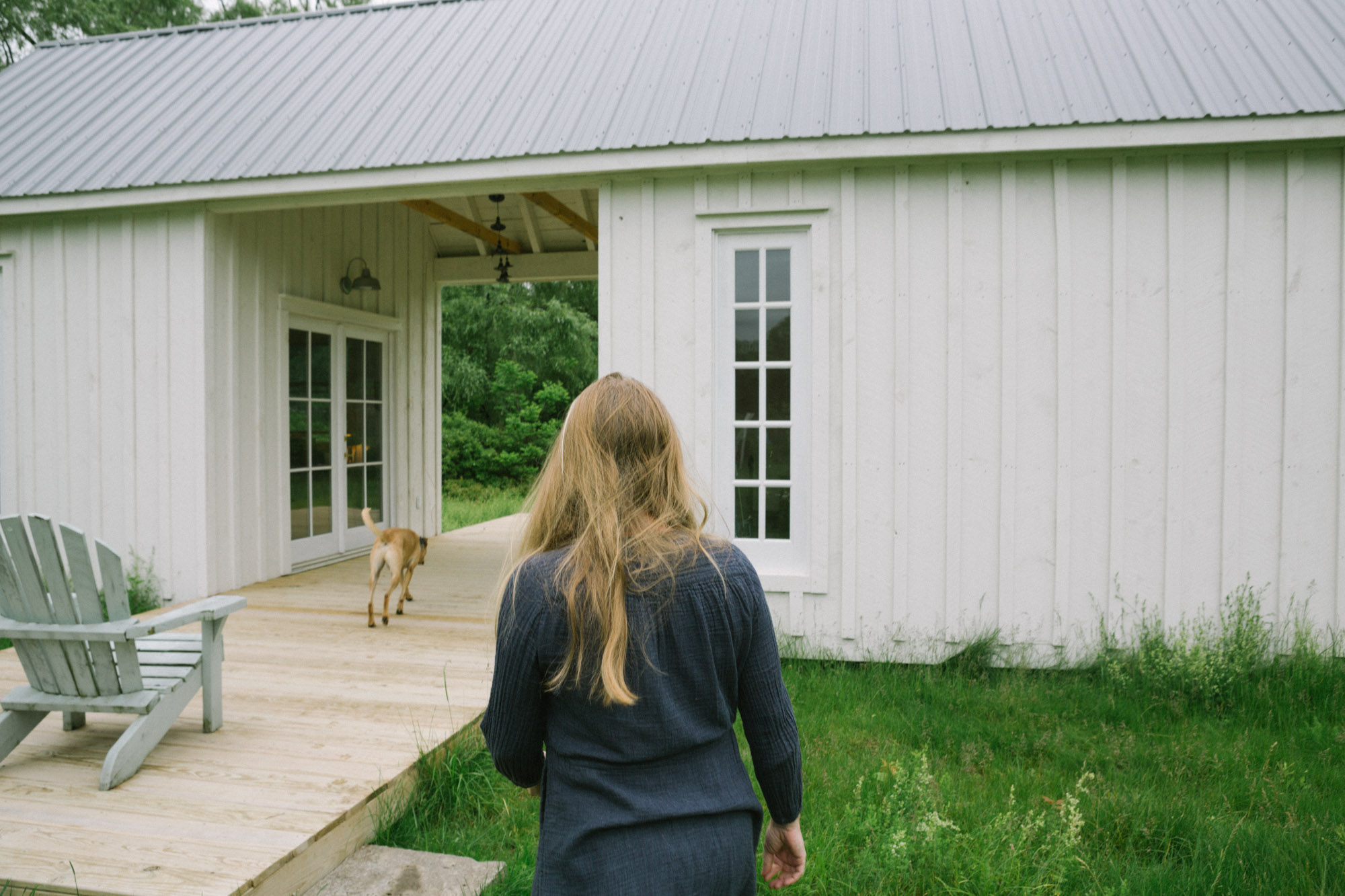 Samantha Hunt walks across a green yard toward a writing studio with a breezeway in the center, where her dog is jogging through