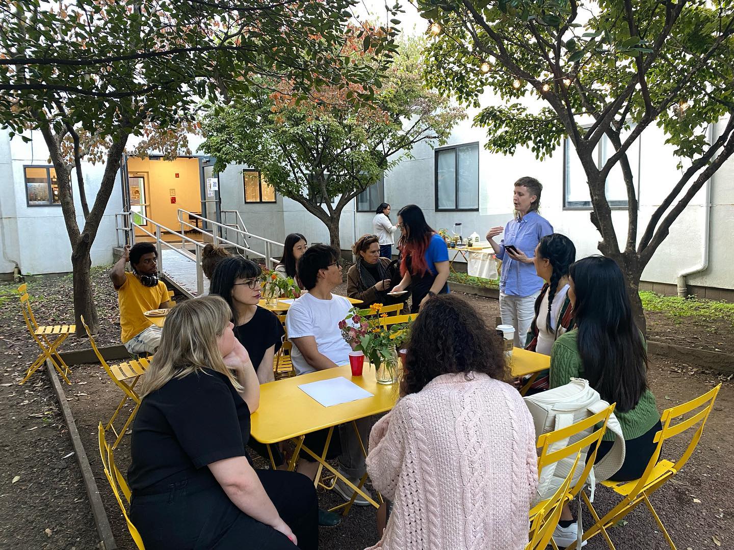 A group of students sitting in garden enclosed by white buildings and surrounded by trees and plants. They are listening to a faculty member.