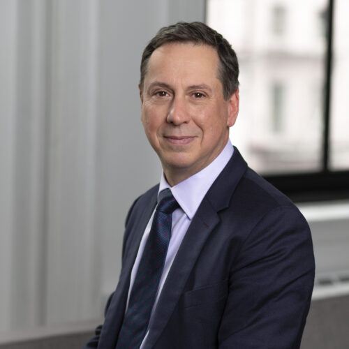 Portrait of a man in a dark suit and tie, sitting in an office environment with a soft smile.