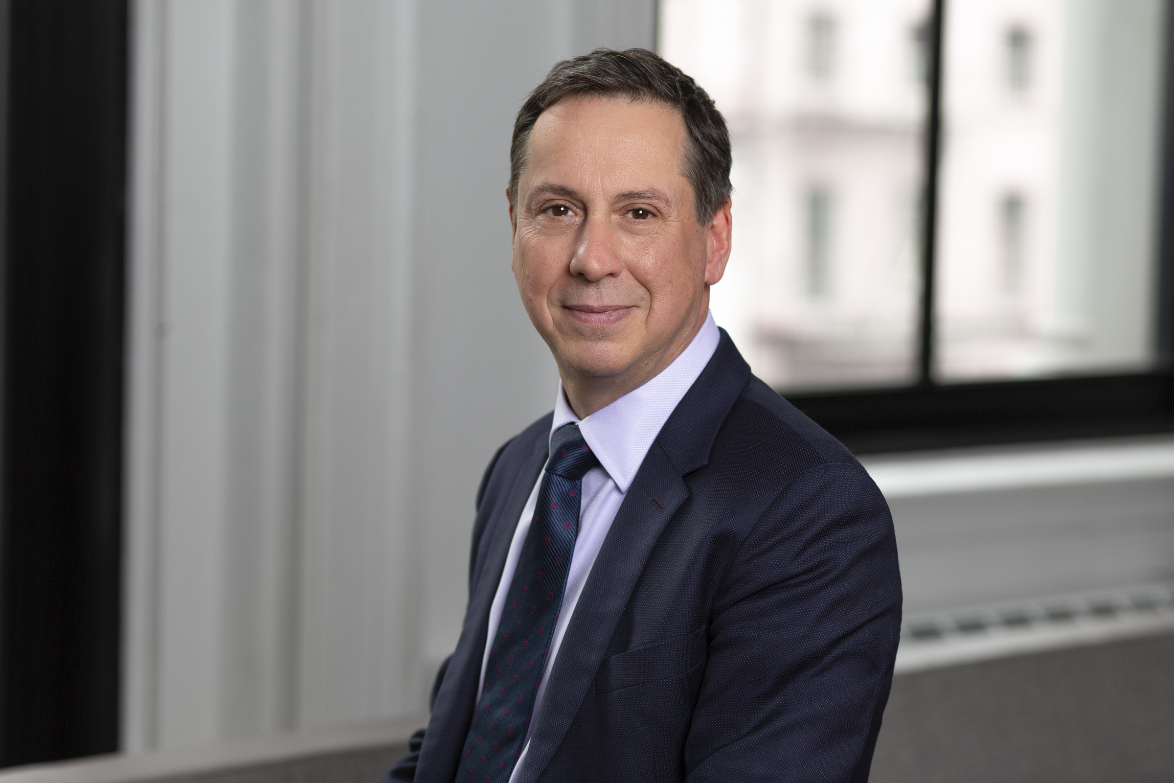 Portrait of a man in a dark suit and tie, sitting in an office environment with a soft smile.