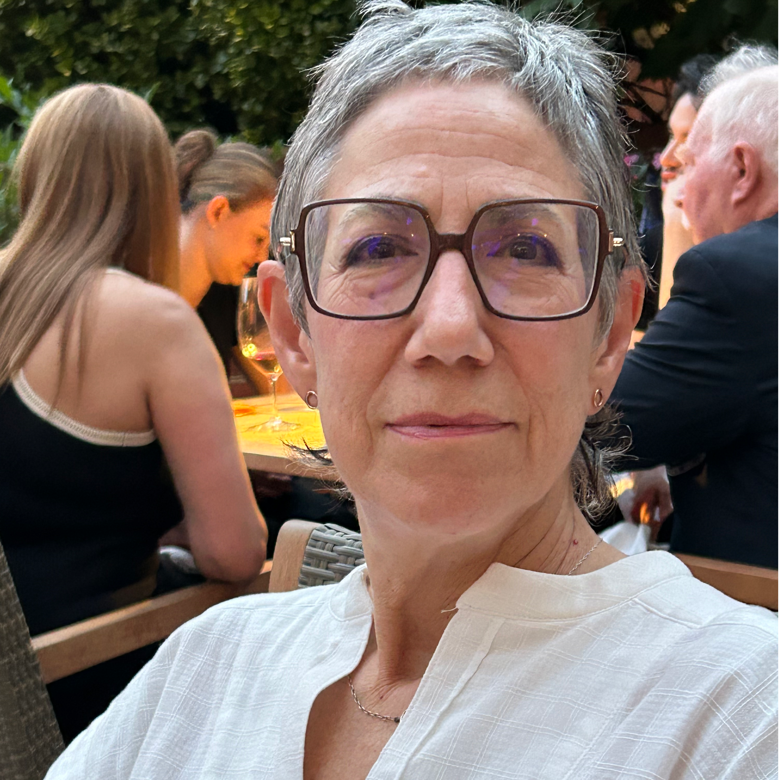 Portrait of an older woman with short gray hair, wearing rectangular glasses and a white linen V-neck shirt. She has a serene expression and a slight smile, seated in what appears to be an outdoor dining area with other guests visible in the background.