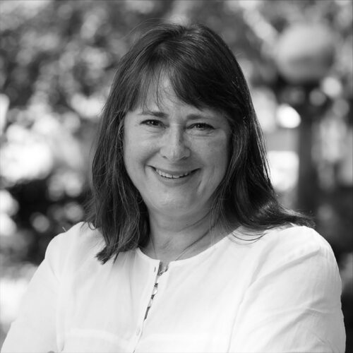 A black-and-white portrait of Rebeccah Pailes-Friedman, smiling warmly in an outdoor setting, wearing a light-colored shirt.