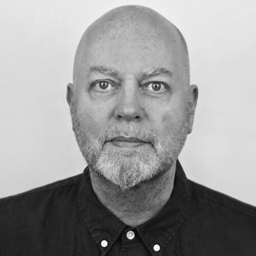 A black-and-white close-up of John-Michael Ekeblad, facing the camera with a neutral expression, wearing a button-up shirt with a short, white beard.