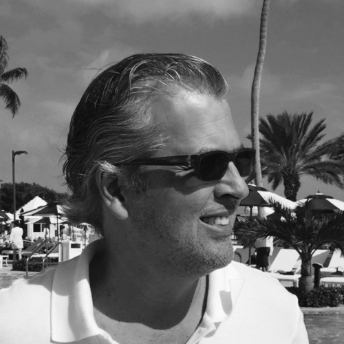 A grayscale side profile of Allen Wilpon, a man with slicked-back hair, wearing sunglasses, smiling outdoors in a sunny location with palm trees in the background.