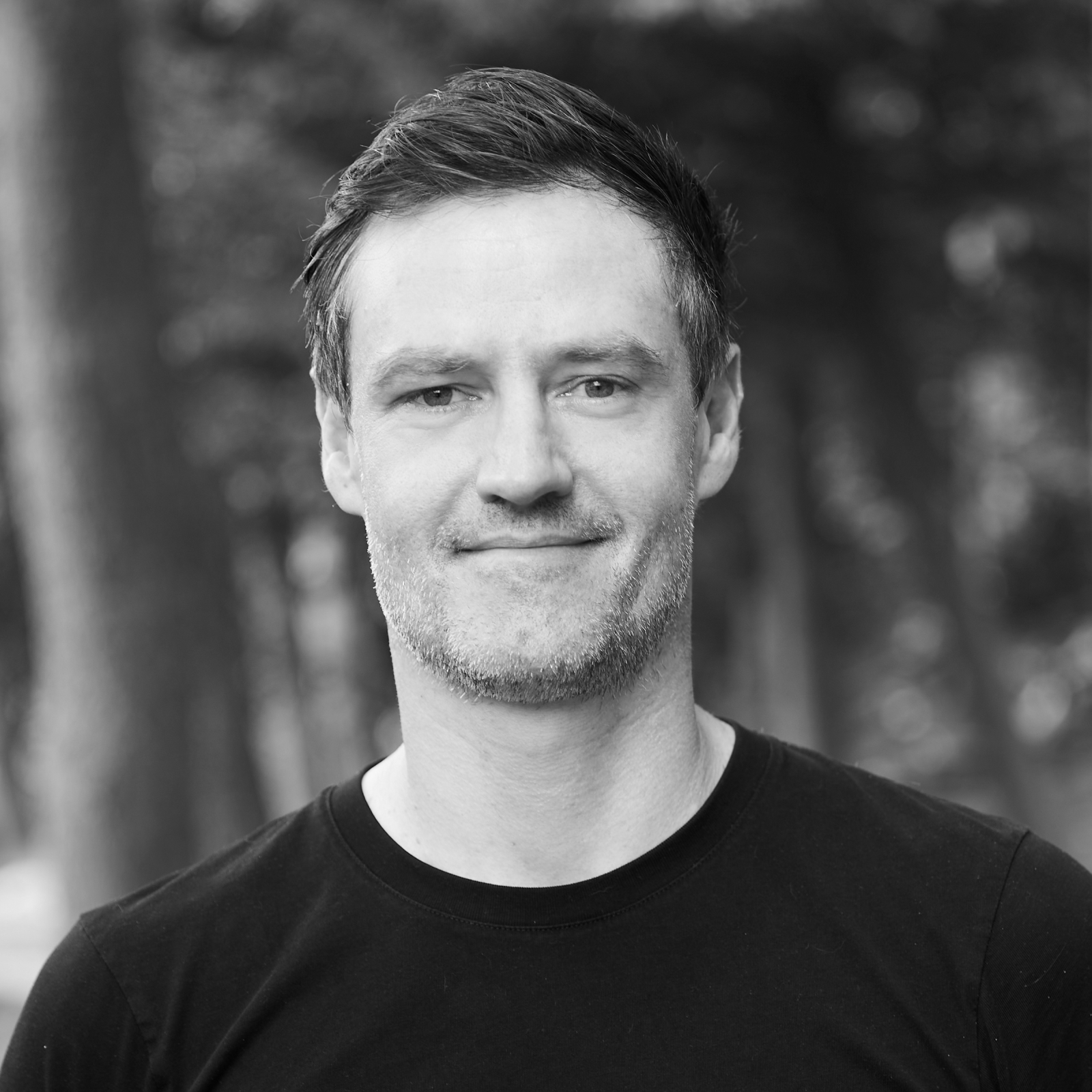 A grayscale portrait of Andre Detller, a man with a close-cropped haircut and beard, wearing a dark turtleneck, looking to the side in a forest setting.