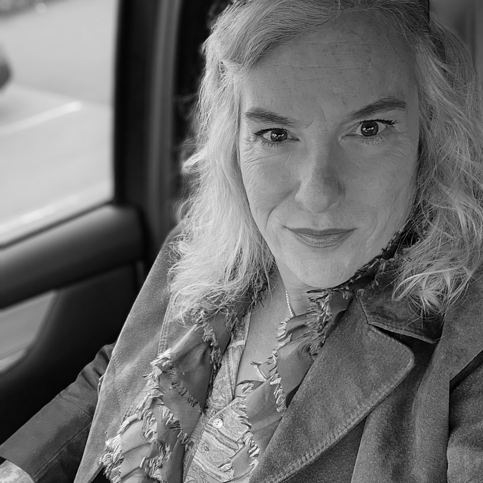 A grayscale portrait of a woman with wavy hair sitting in a car. She is looking directly at the camera with a confident expression. She is wearing a scarf and a jacket, and natural light is coming in through the car window.