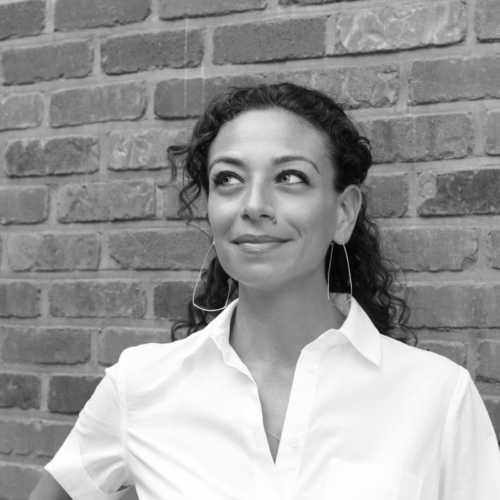 A black and white portrait of a woman with curly hair, standing against a brick wall, looking off to the side with a slight smile.